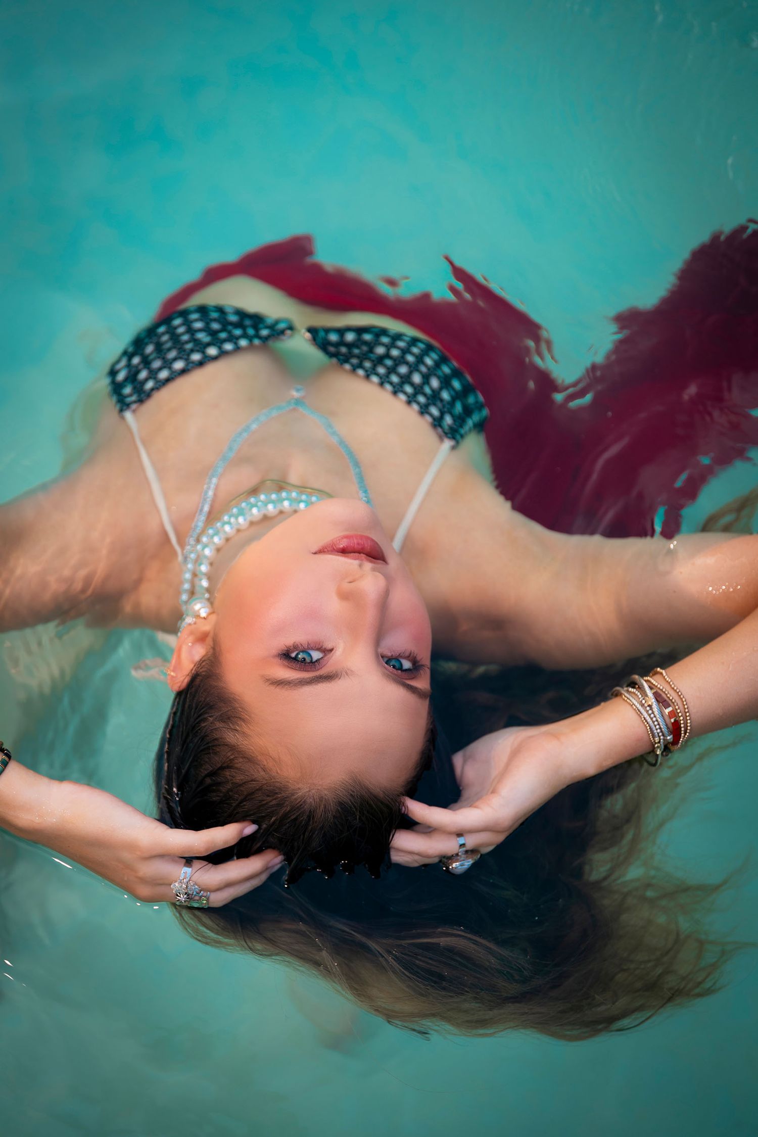 Woman floating in turquoise pool wearing patterned bikini top with pearl headpiece and burgundy skirt, hair spread in water.