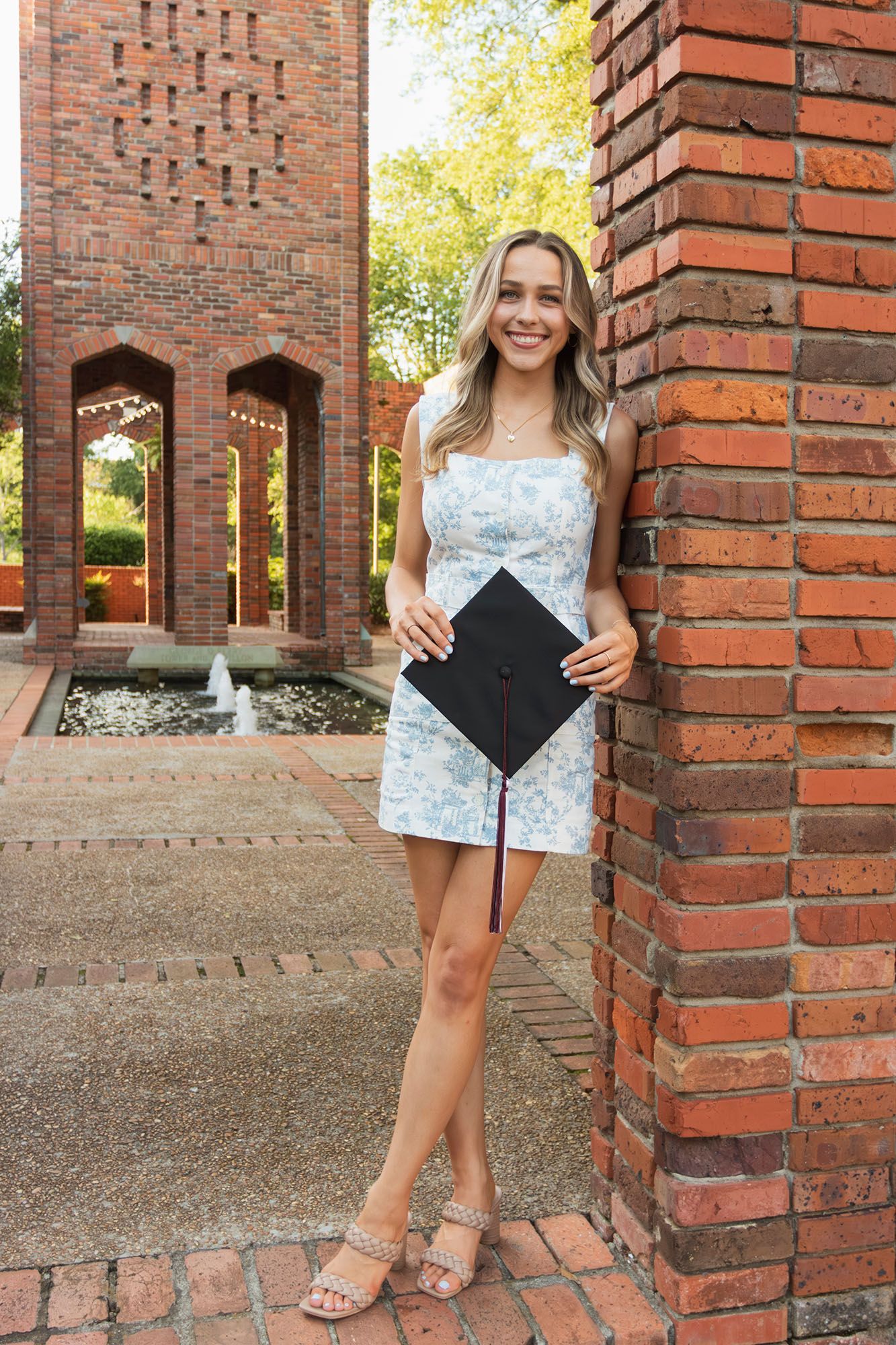 Blonde female graduate in floral dress holds cap while leaning against brick wall near fountain.
