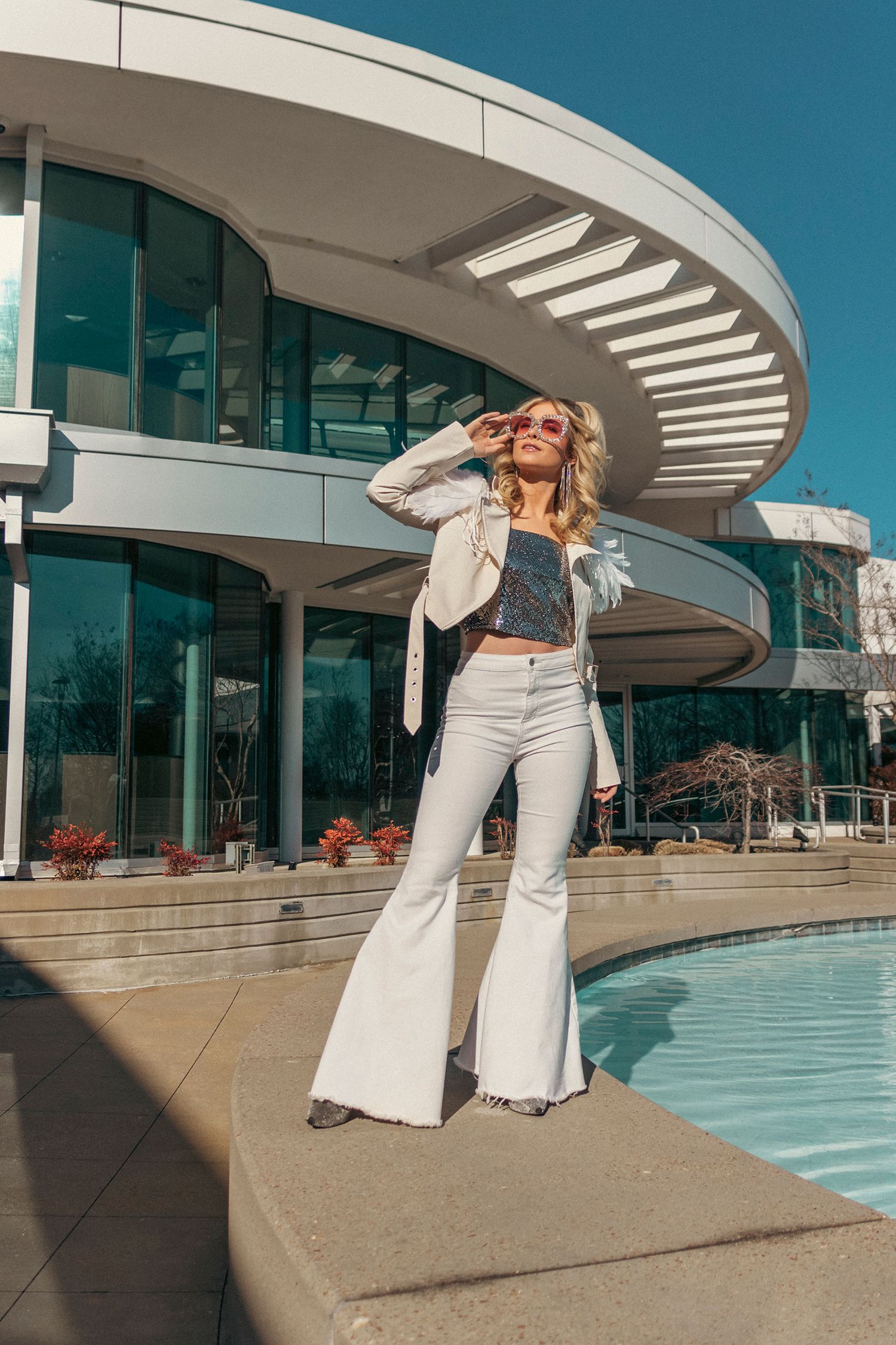 Blonde woman in white flare jeans and crop top poses by modern circular building poolside on sunny day.