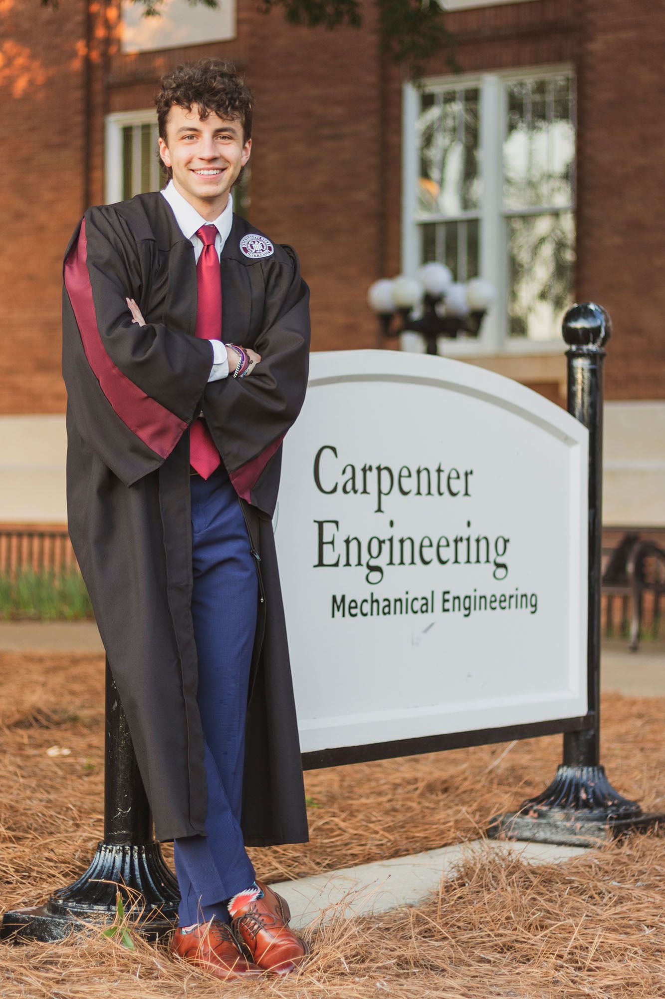 Smiling male graduate in black robe and red tie poses beside Carpenter Mechanical Engineering sign.