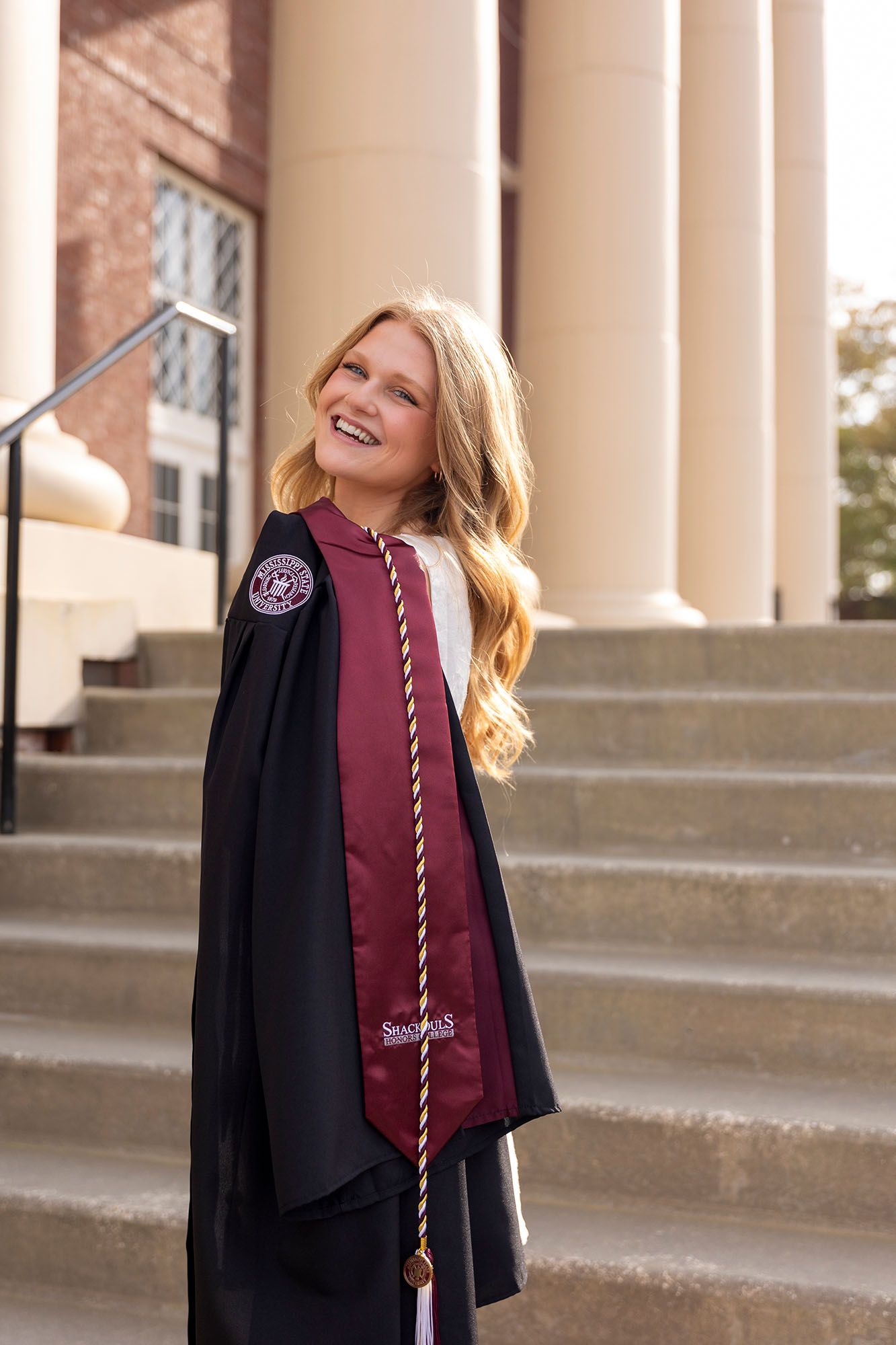 Happy female graduate in black gown and maroon stole poses on university building steps.