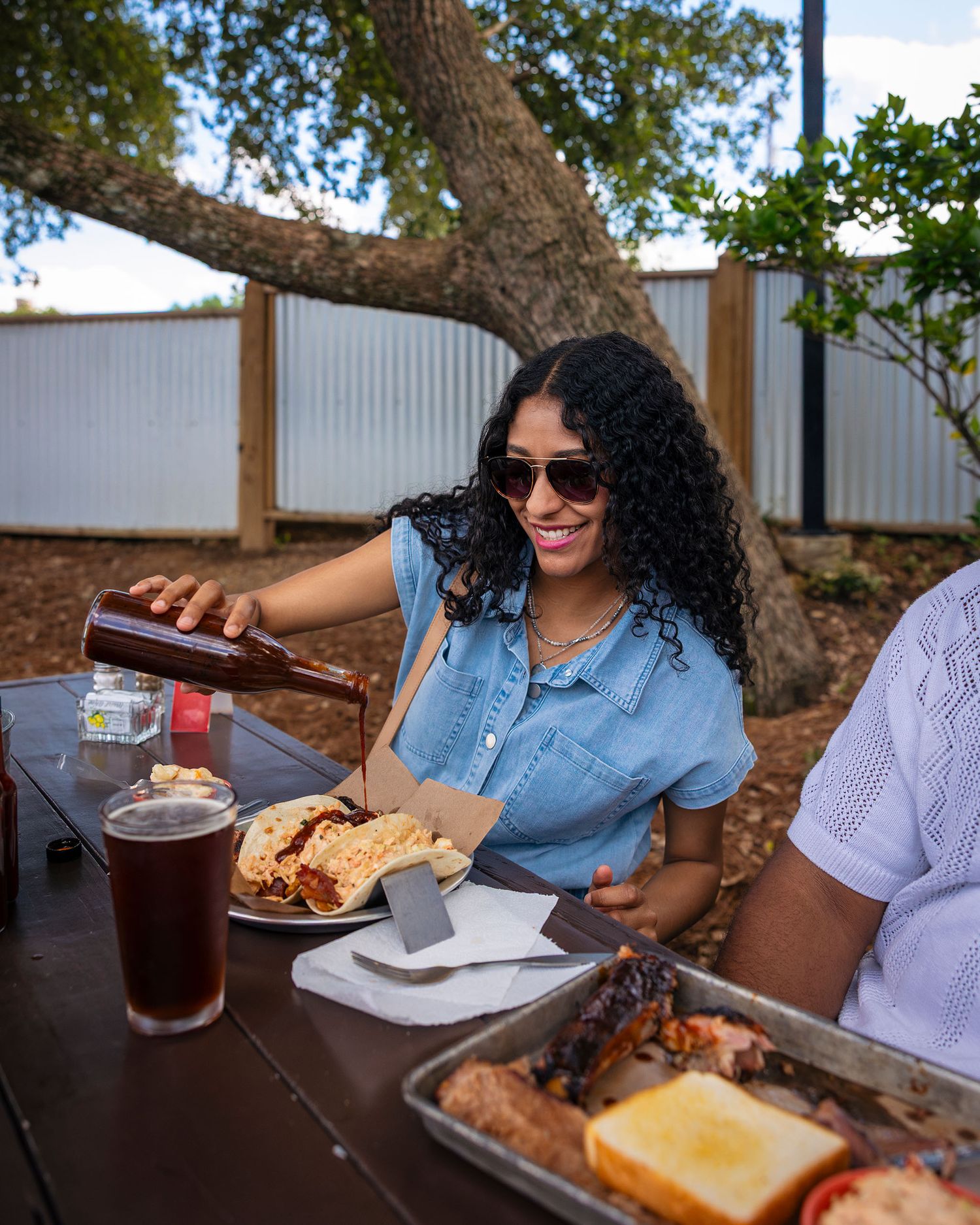 A woman pouring Kona beer at an outdoor BBQ table with smoked meats and sides on a sunny day.