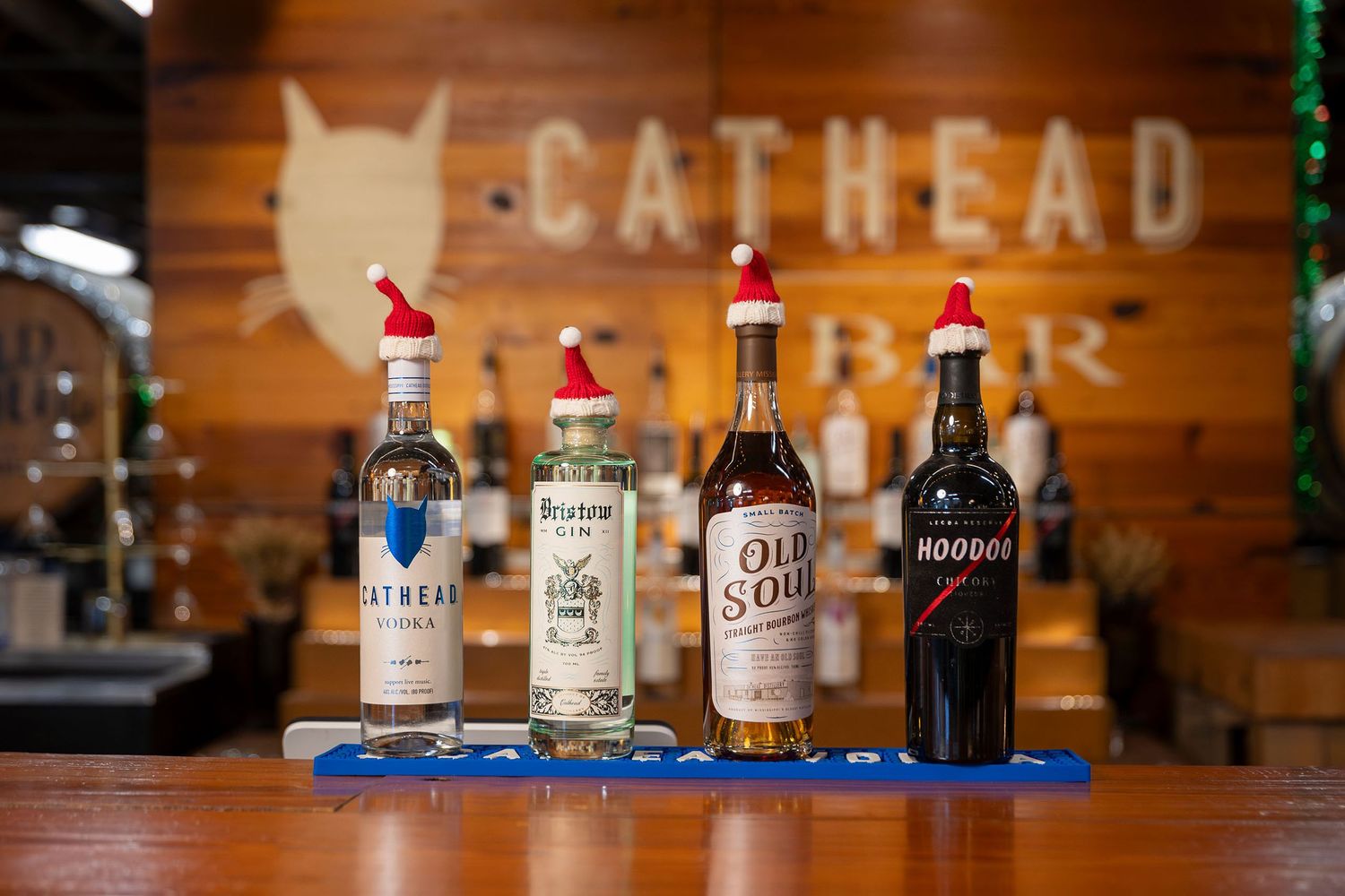 Four Cathead Distillery spirit bottles wearing Santa hats displayed on a wooden bar.