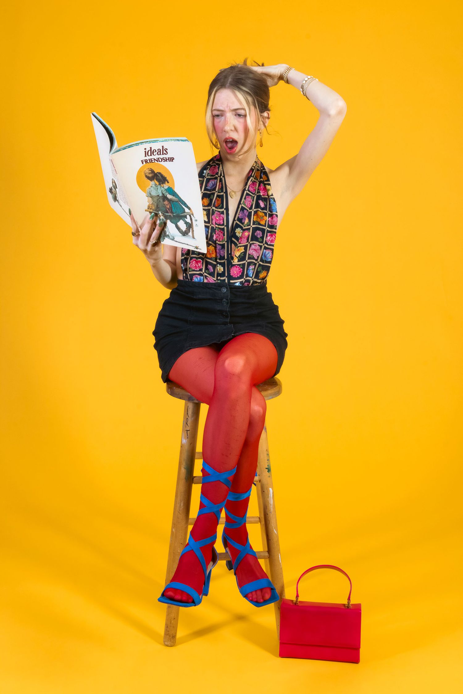 Blonde woman in colorful outfit sits on stool reading a magazine on yellow background.