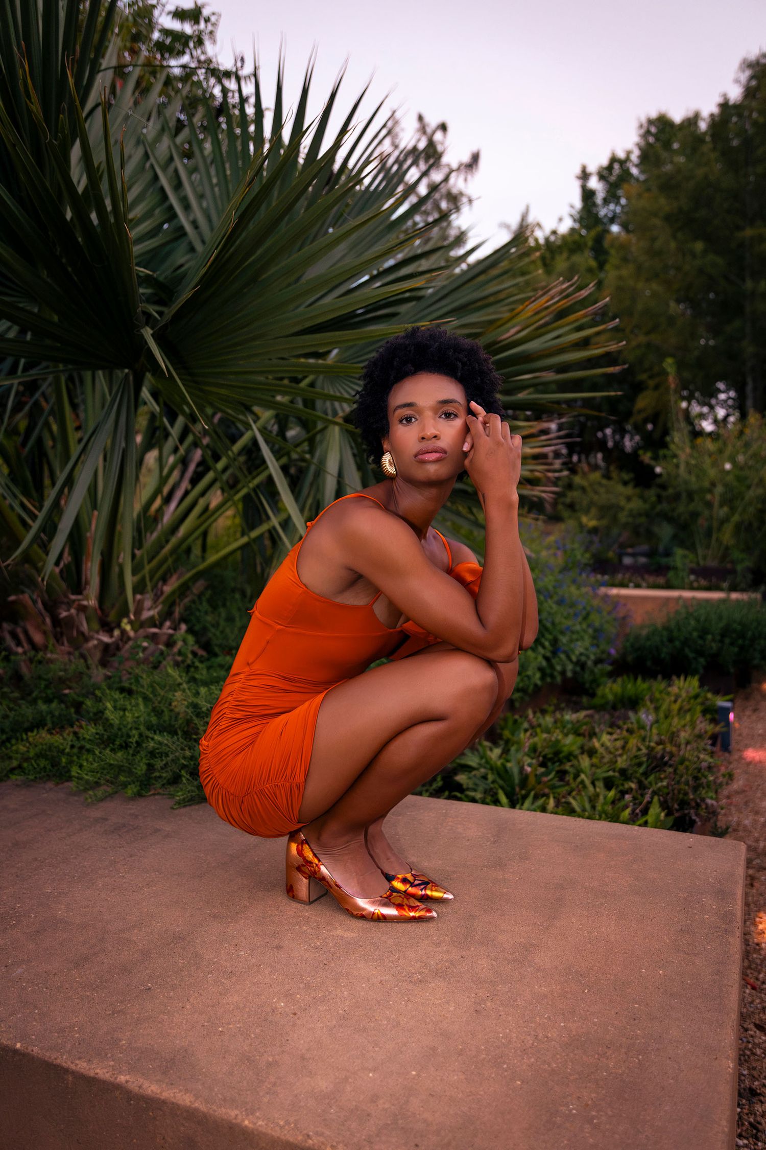 Woman in orange ruched mini dress crouching on stone ledge with tropical palm backdrop.