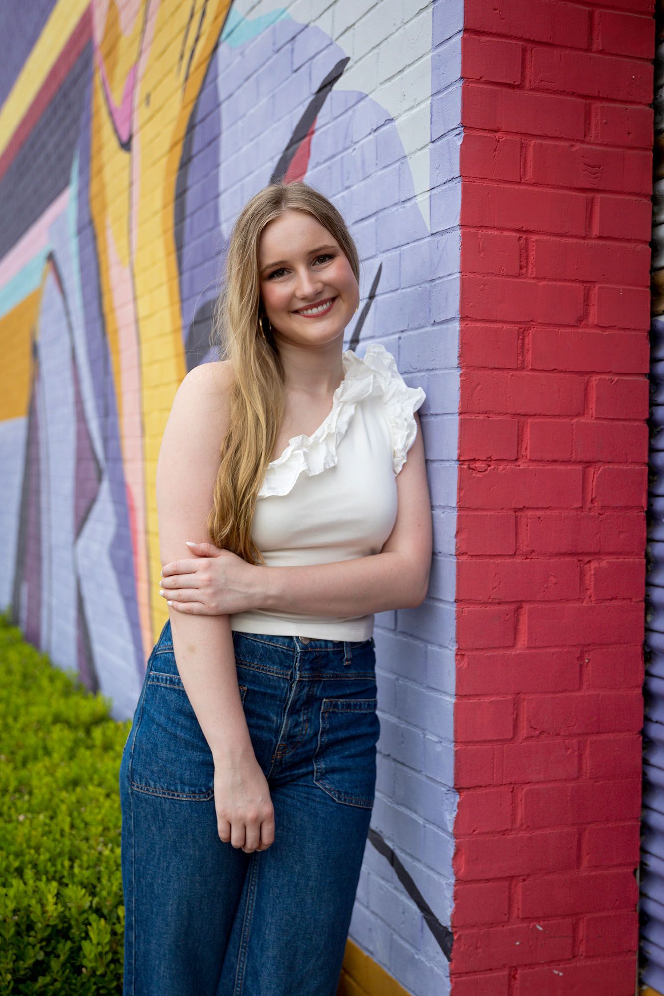 Smiling young woman in white ruffled top and jeans poses beside a vibrant painted brick wall.