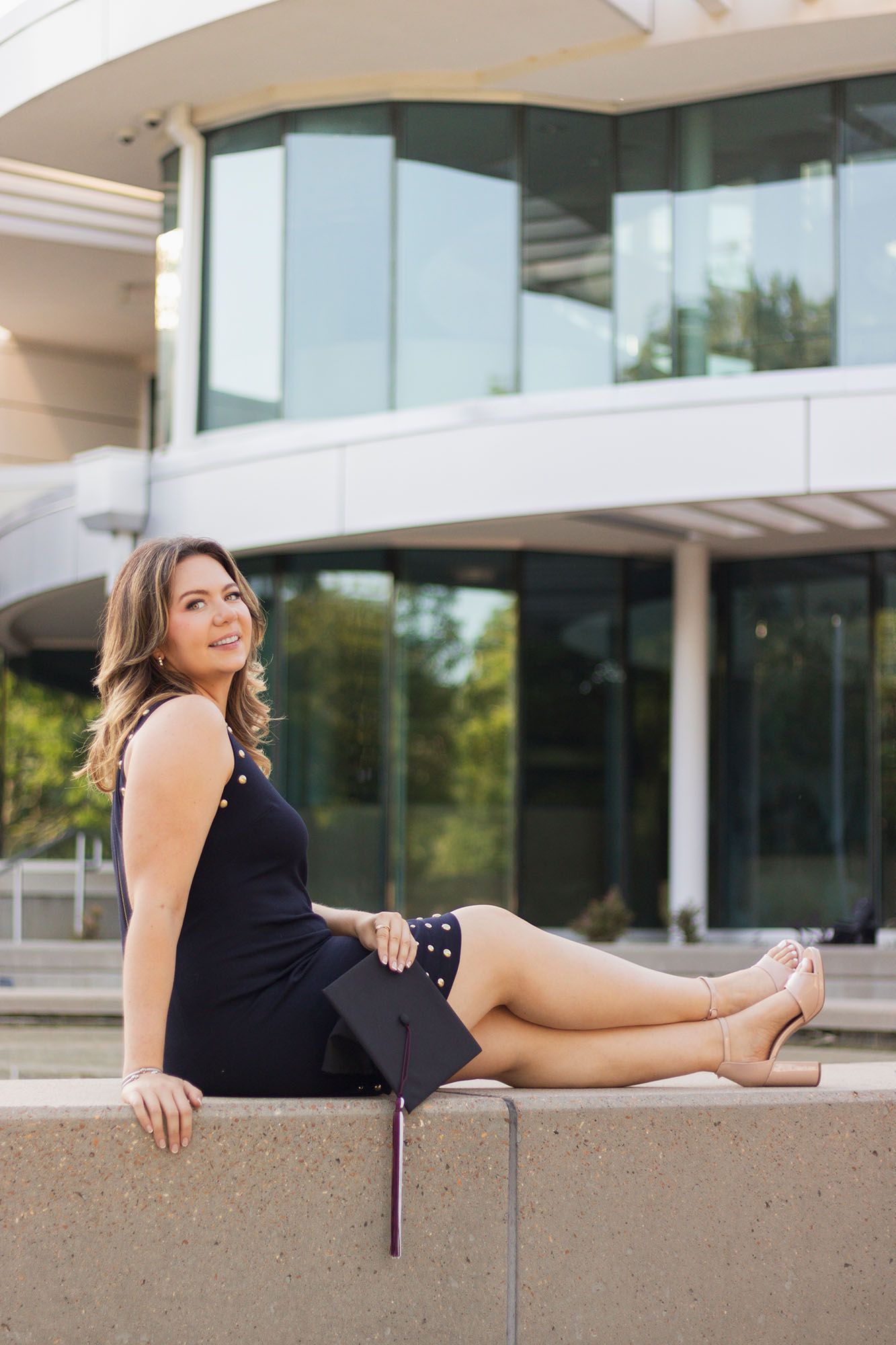 Female graduate in black dress sits on ledge holding cap outside modern university building.