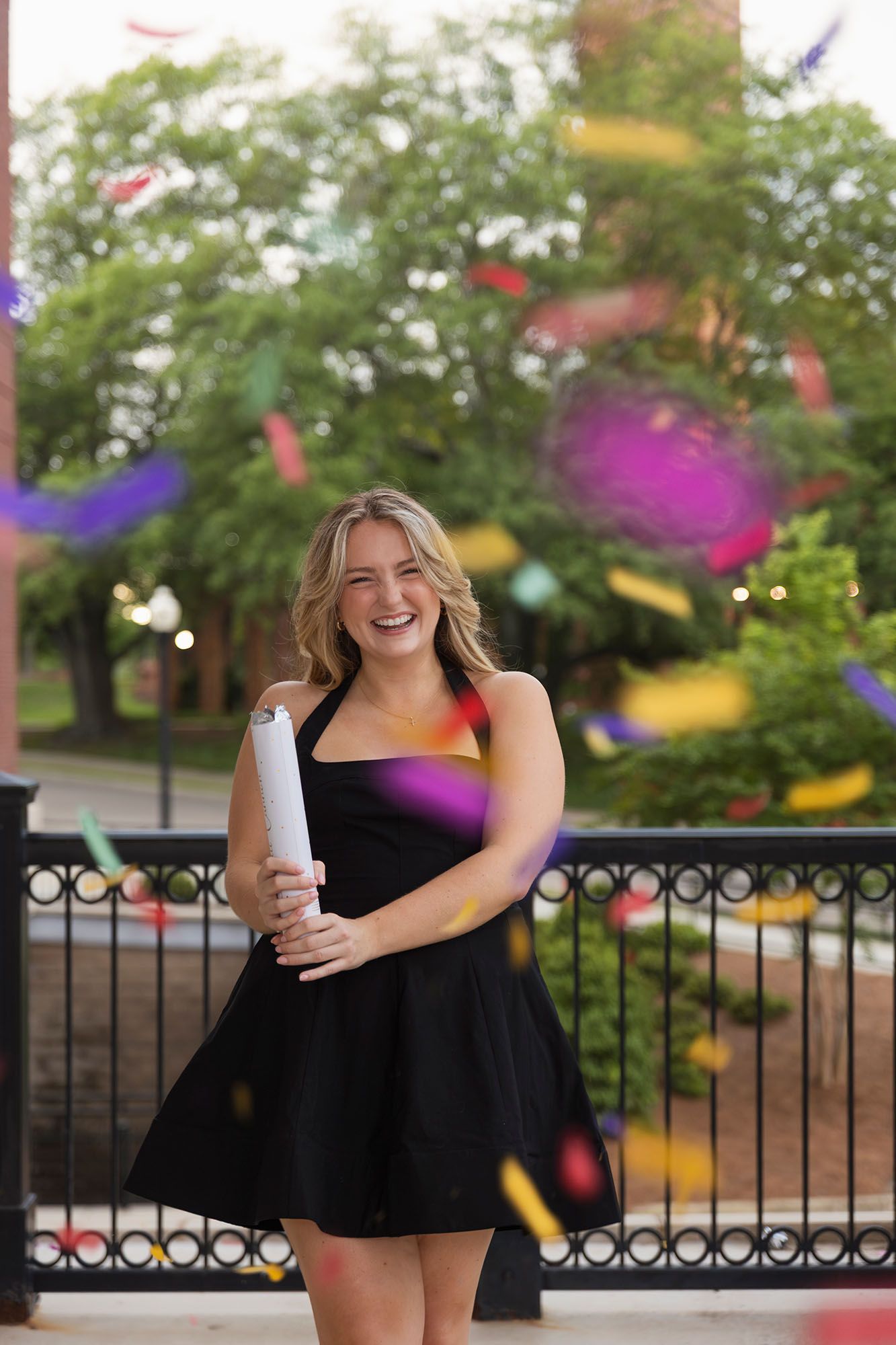 Laughing female graduate in black dress holds diploma scroll amid colorful confetti outdoors.