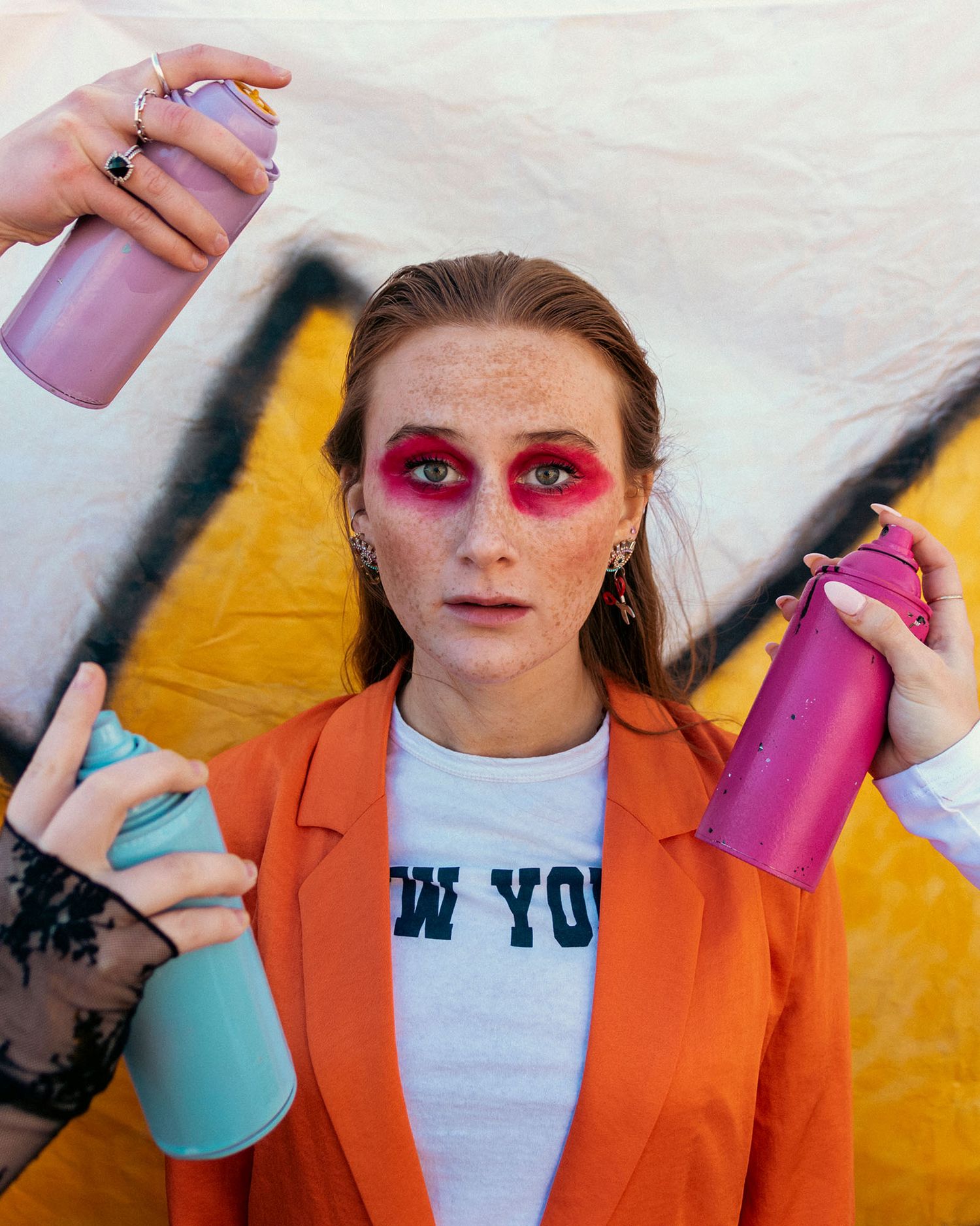 Freckled woman in orange blazer holds colorful spray cans with bold red eye makeup before graffiti wall.