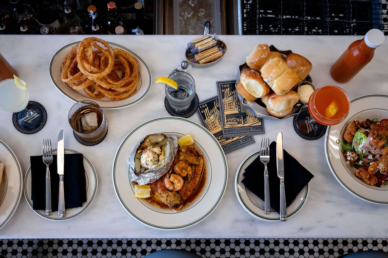 Overhead view of a restaurant table with shrimp dishes, onion rings, rolls, and cocktails.