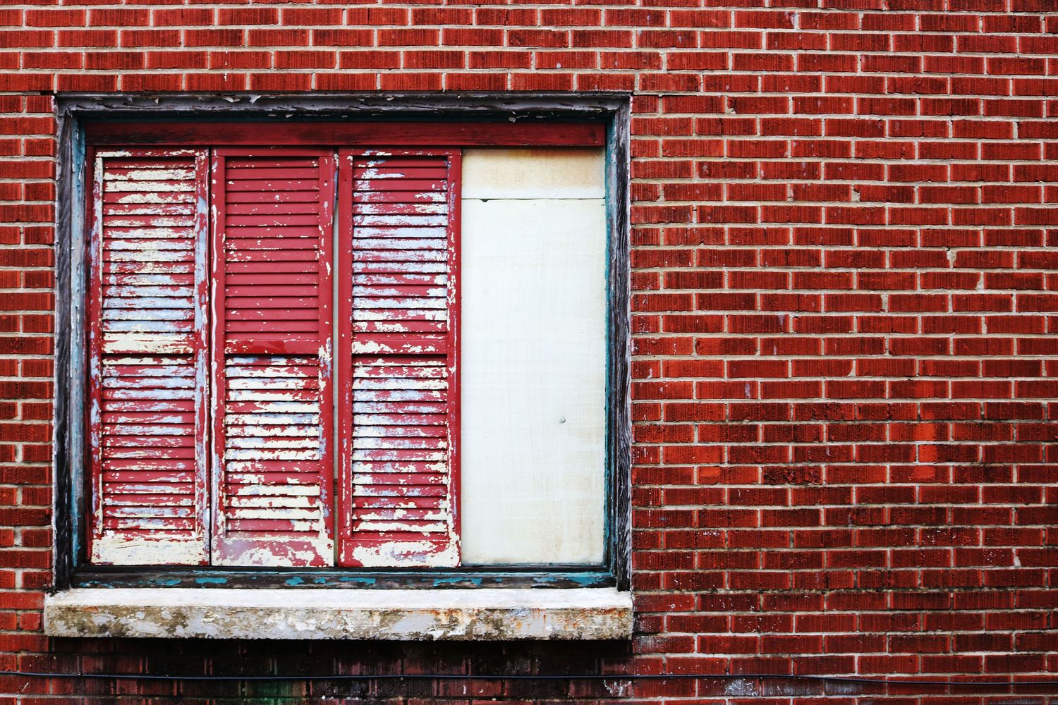 A weathered red brick building with peeling red shutters and a boarded window showing aged urban decay.