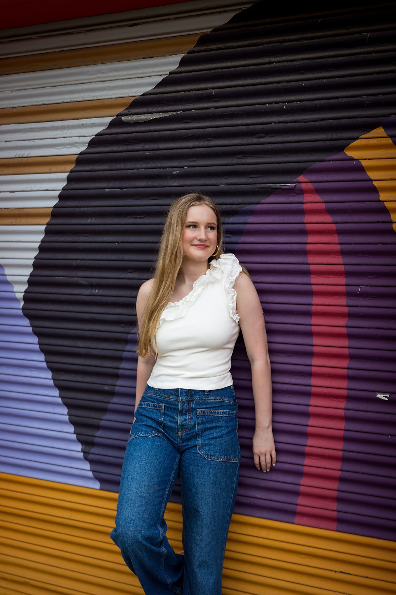 Young blonde woman in white top and jeans leans against a colorful striped mural wall.