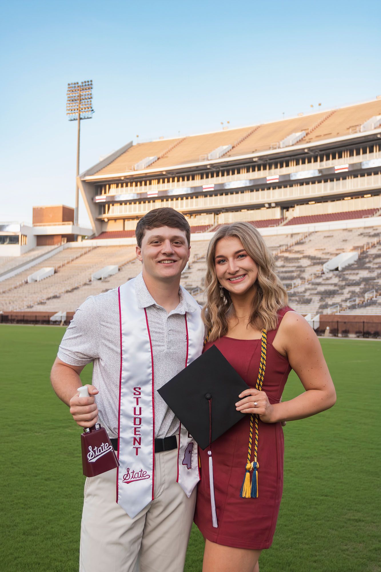 Two Mississippi State graduates smile together on football field with stadium behind them.