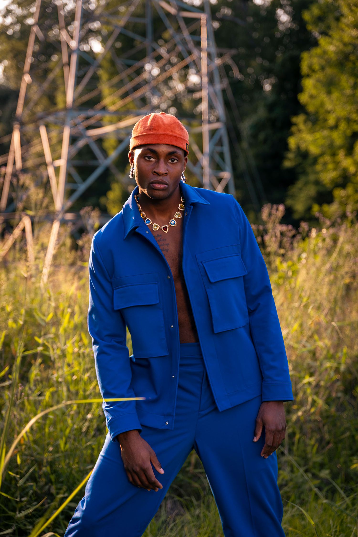 Fashionable man in royal blue suit and orange cap posing in lush green outdoor setting.