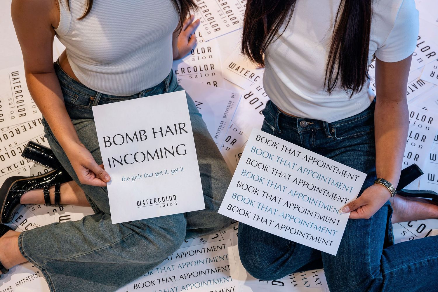 Two women holding signs reading Bomb Hair Incoming and Book That Appointment at Watercolor Salon.