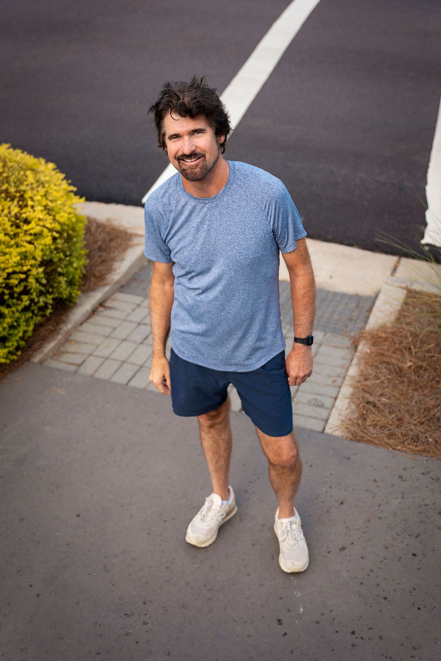 A cheerful bearded man in a blue t-shirt and navy shorts stands on a suburban sidewalk.