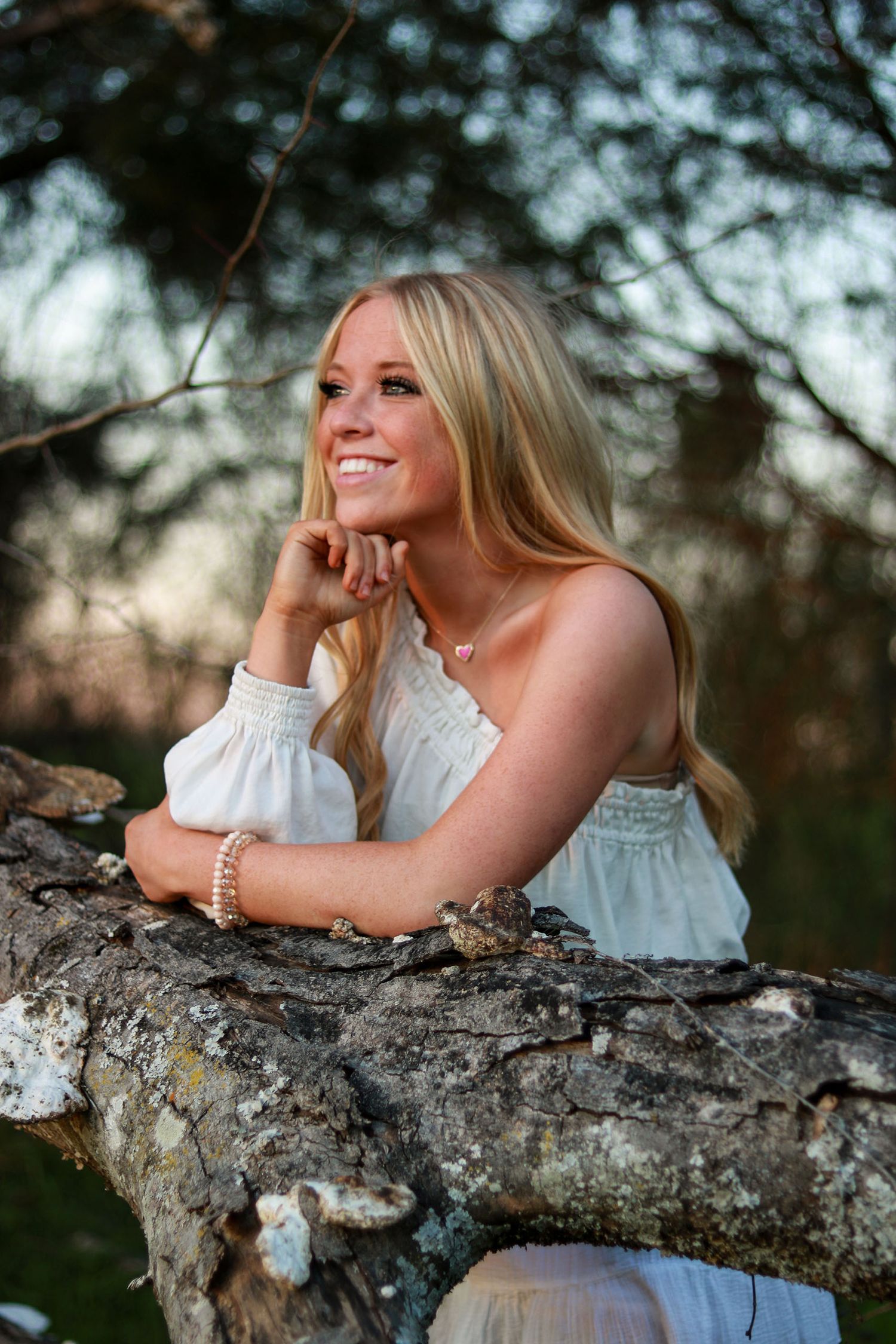 Smiling blonde woman leaning on a tree branch outdoors in a white top during golden hour.
