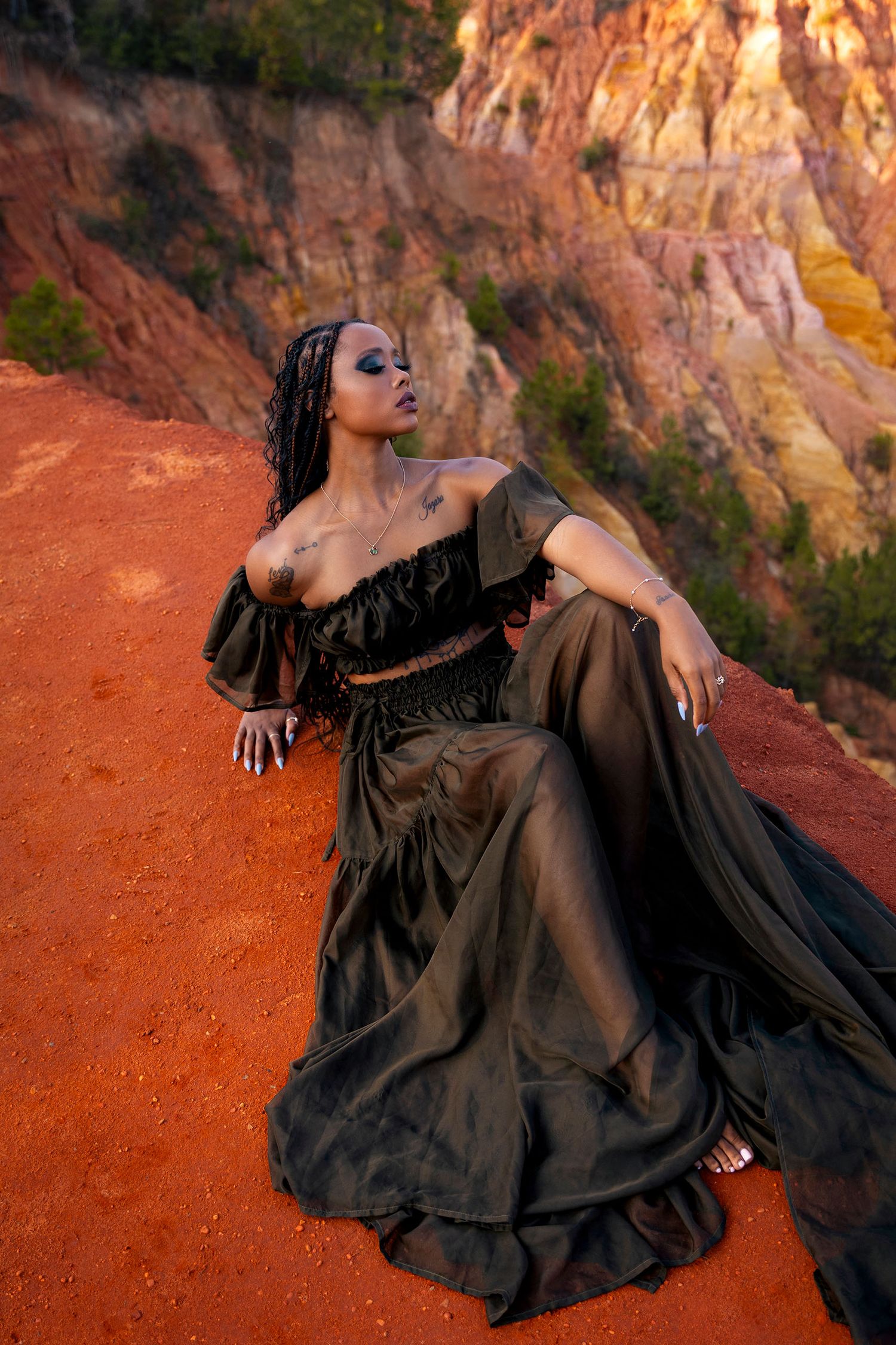 Woman in black off-shoulder crop top and sheer skirt posing on red clay canyon terrain.