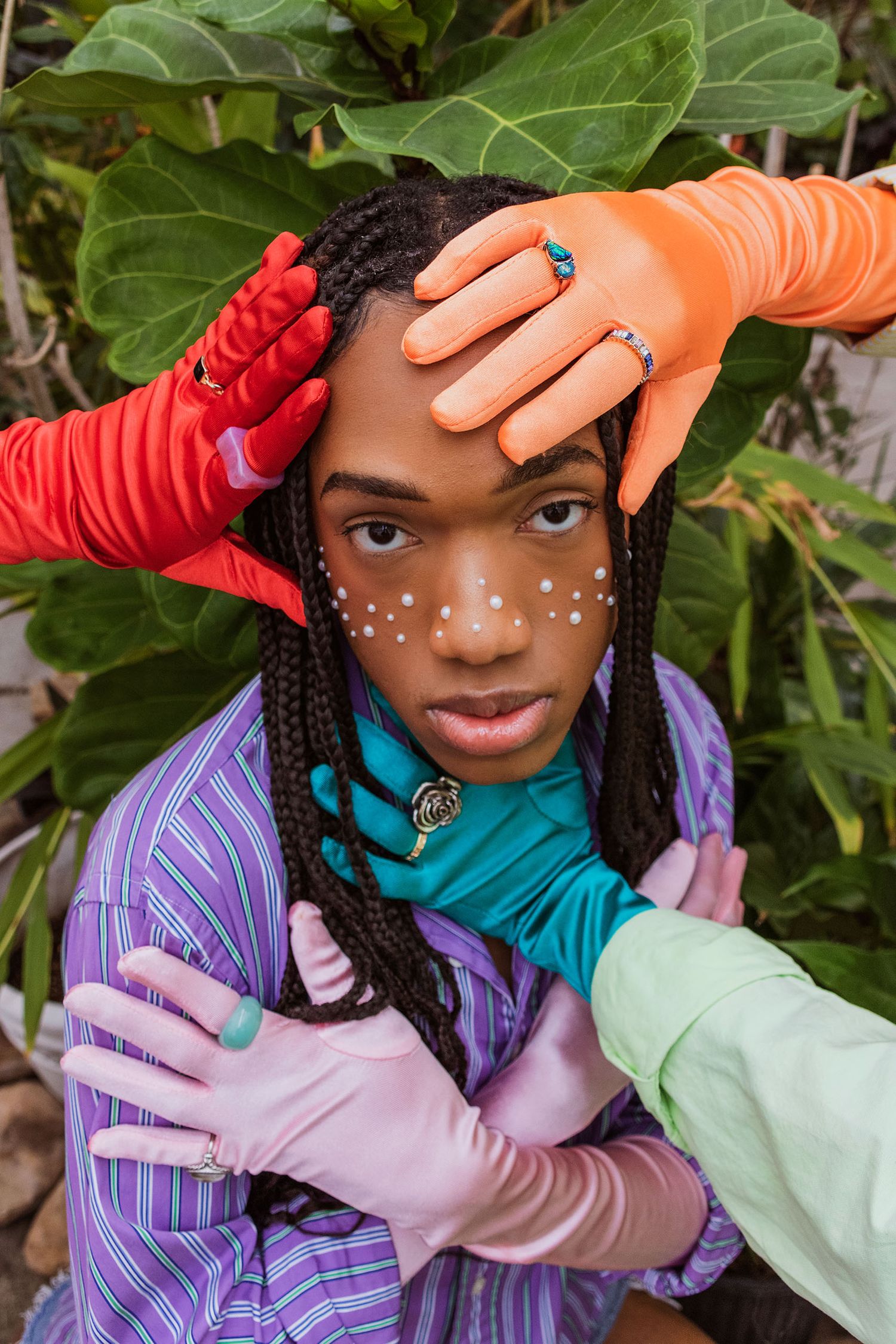 Teen with braids and face dots surrounded by colorful gloved hands amid tropical leaves.