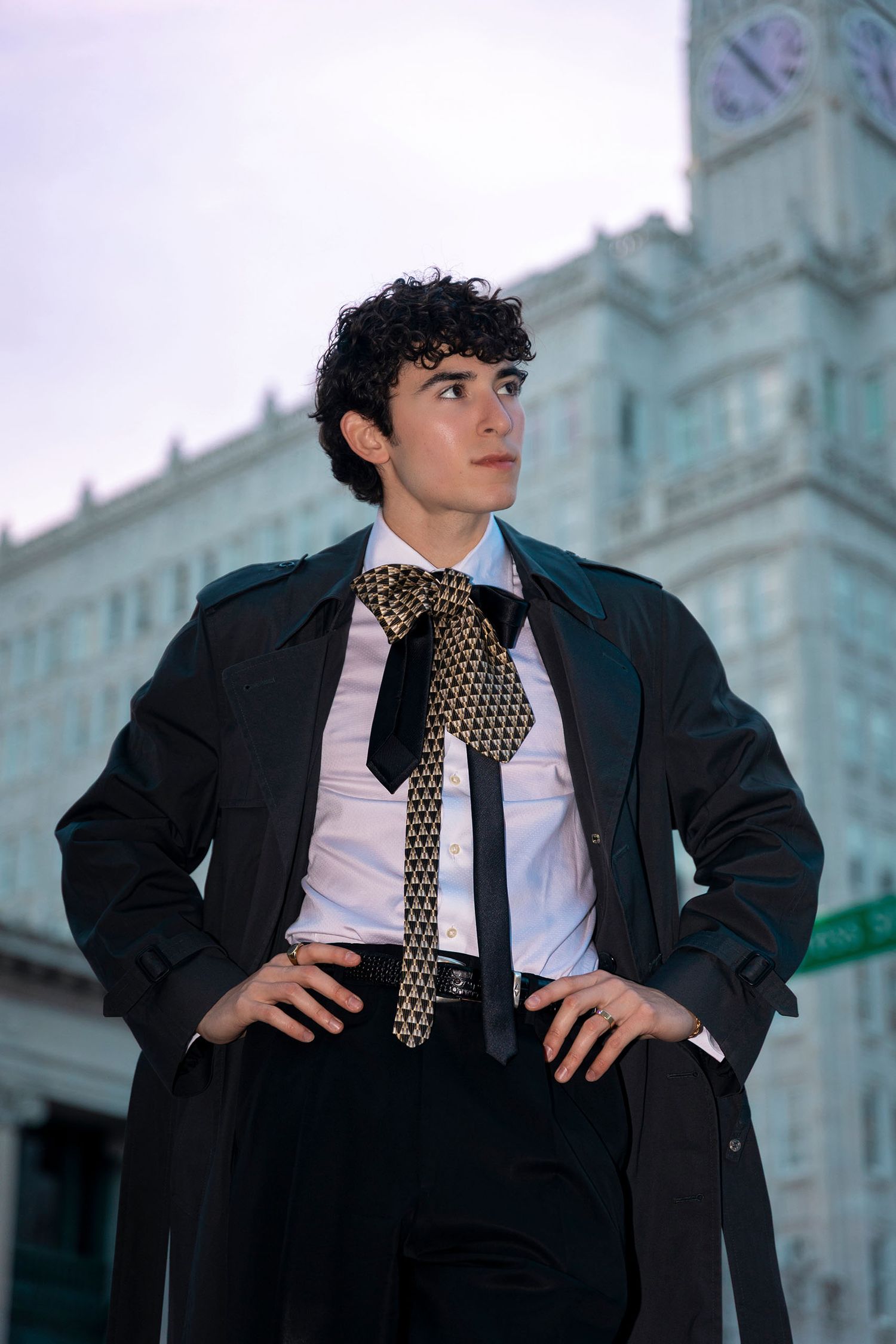 Young man in black trench coat with gold patterned bow tie posing confidently outdoors near historic building.