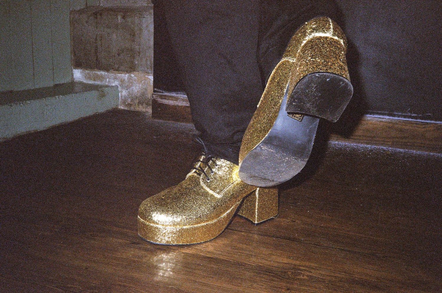 Sparkly gold glitter platform shoes resting on a wooden floor near a fireplace.