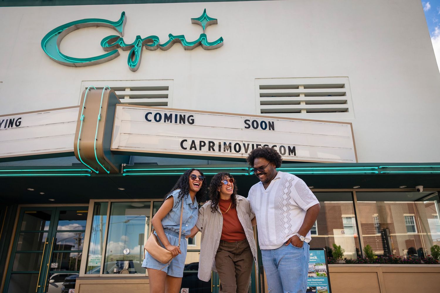 Three friends pose outside the Capri Theater marquee reading Coming Soon in a sunny urban setting.