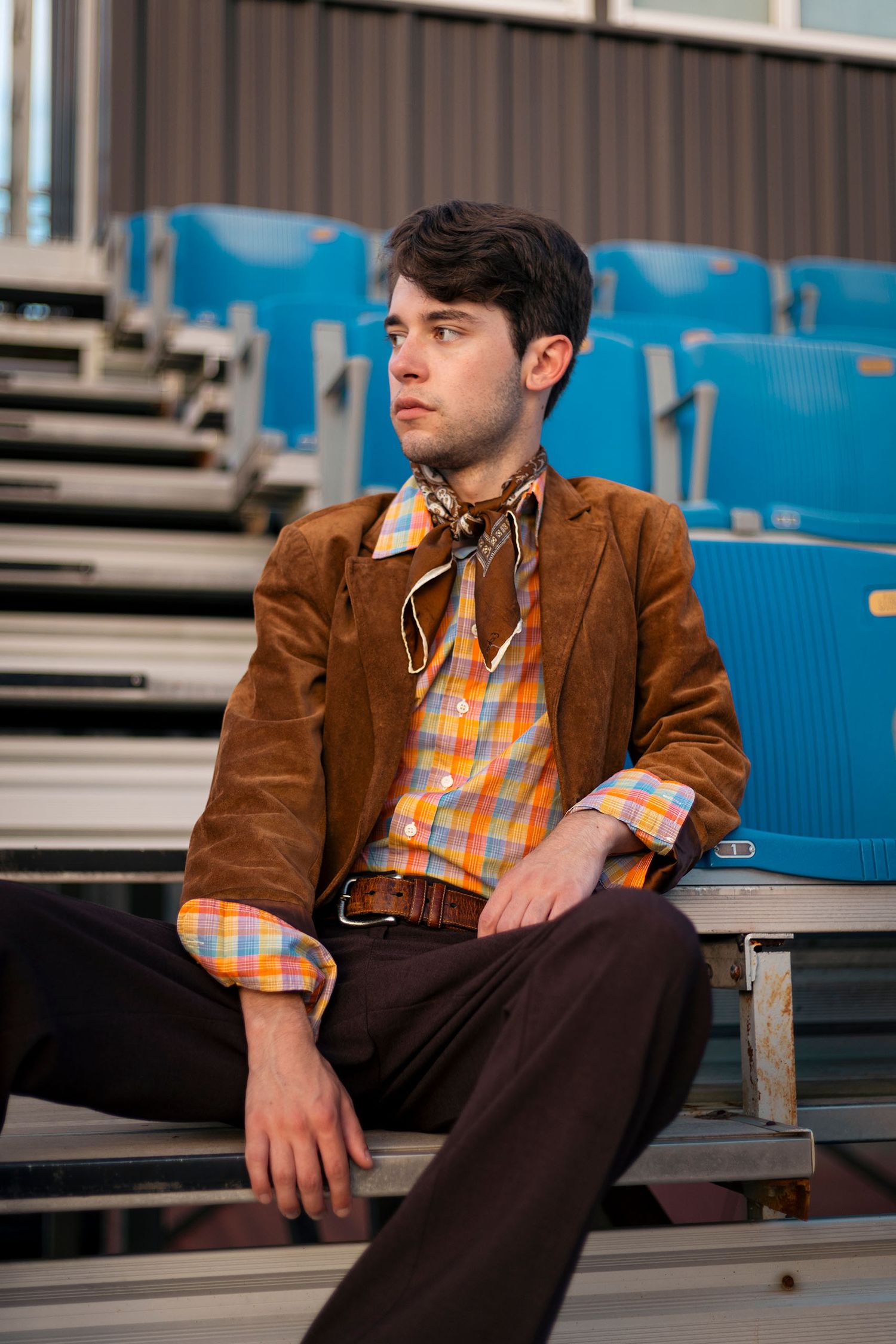 Man in brown suede blazer and plaid shirt sits on stadium bleachers with neck scarf.