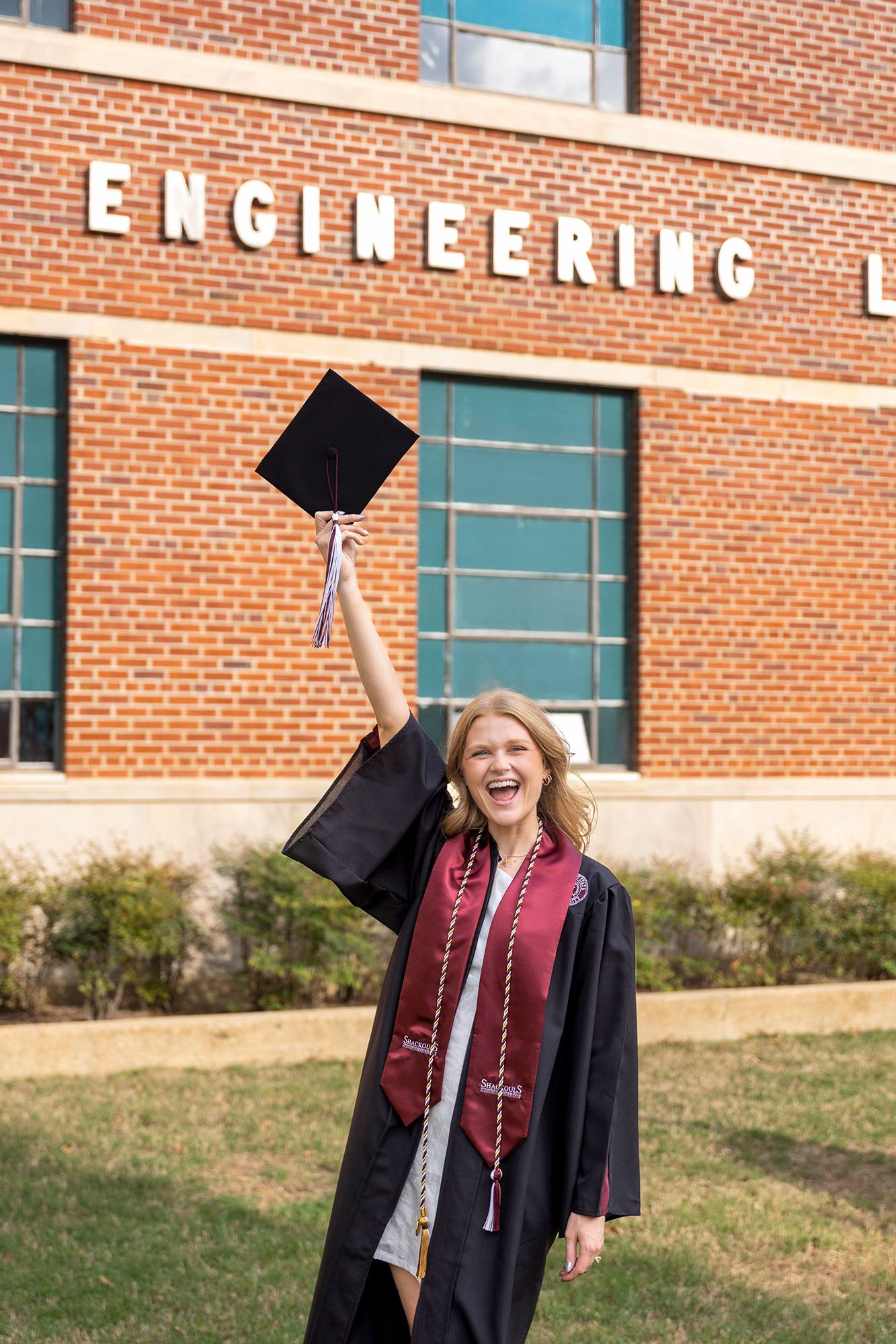 Excited female graduate raises cap in front of Engineering building in black gown.