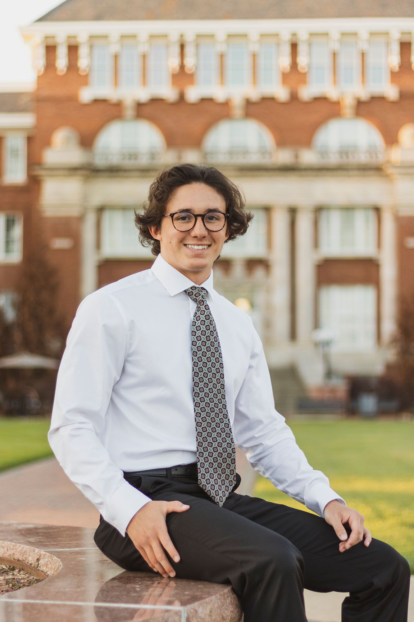 Smiling young man in white dress shirt and tie sitting outdoors in front of red brick university building.