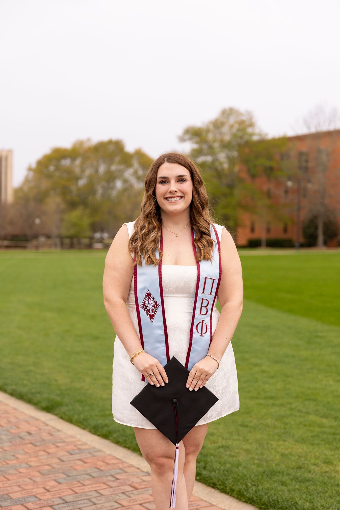 Smiling graduate in white dress wearing Pi Beta Phi stole holding cap on brick campus path.