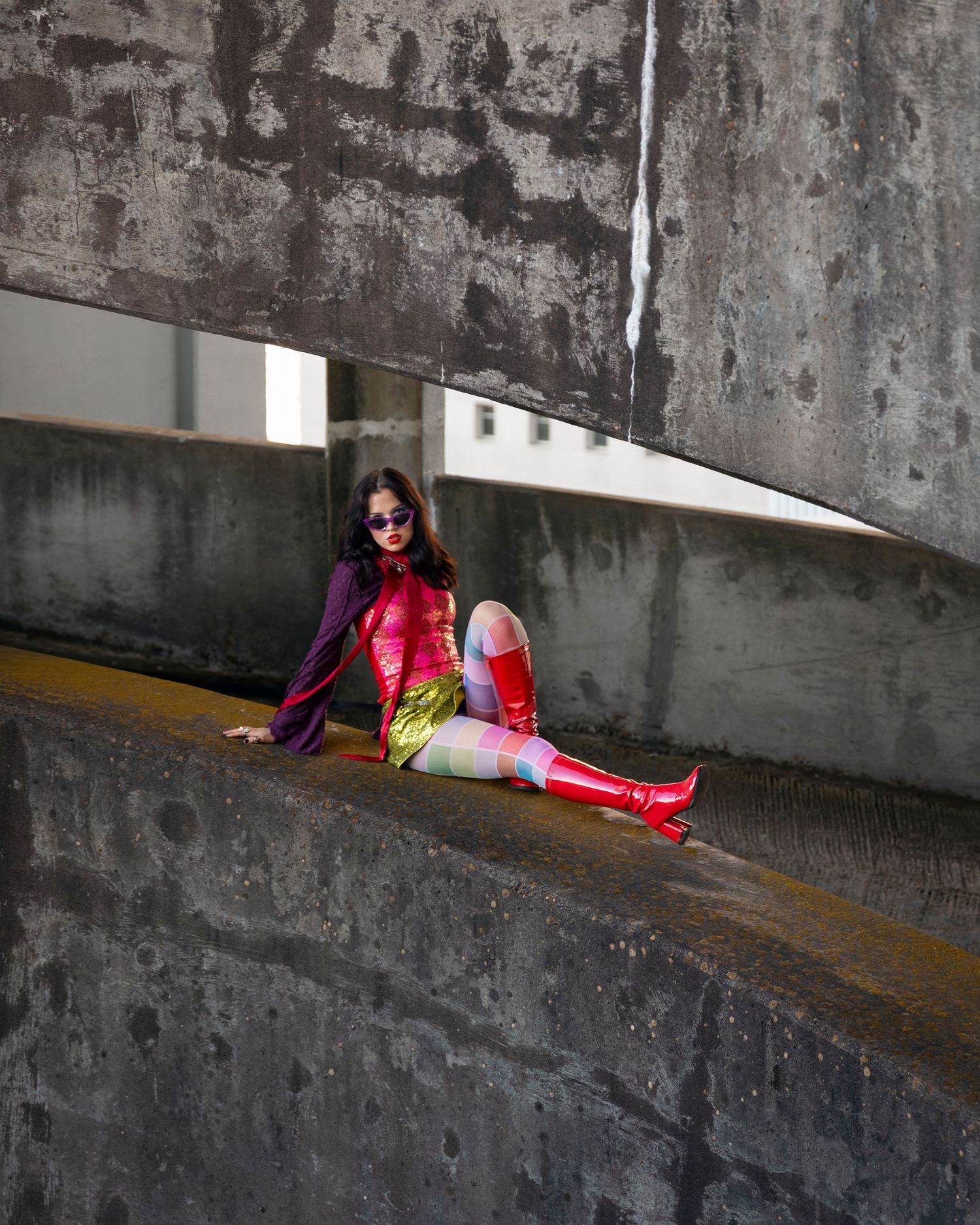 Woman in colorful superhero costume with red boots sitting on urban concrete ledge.
