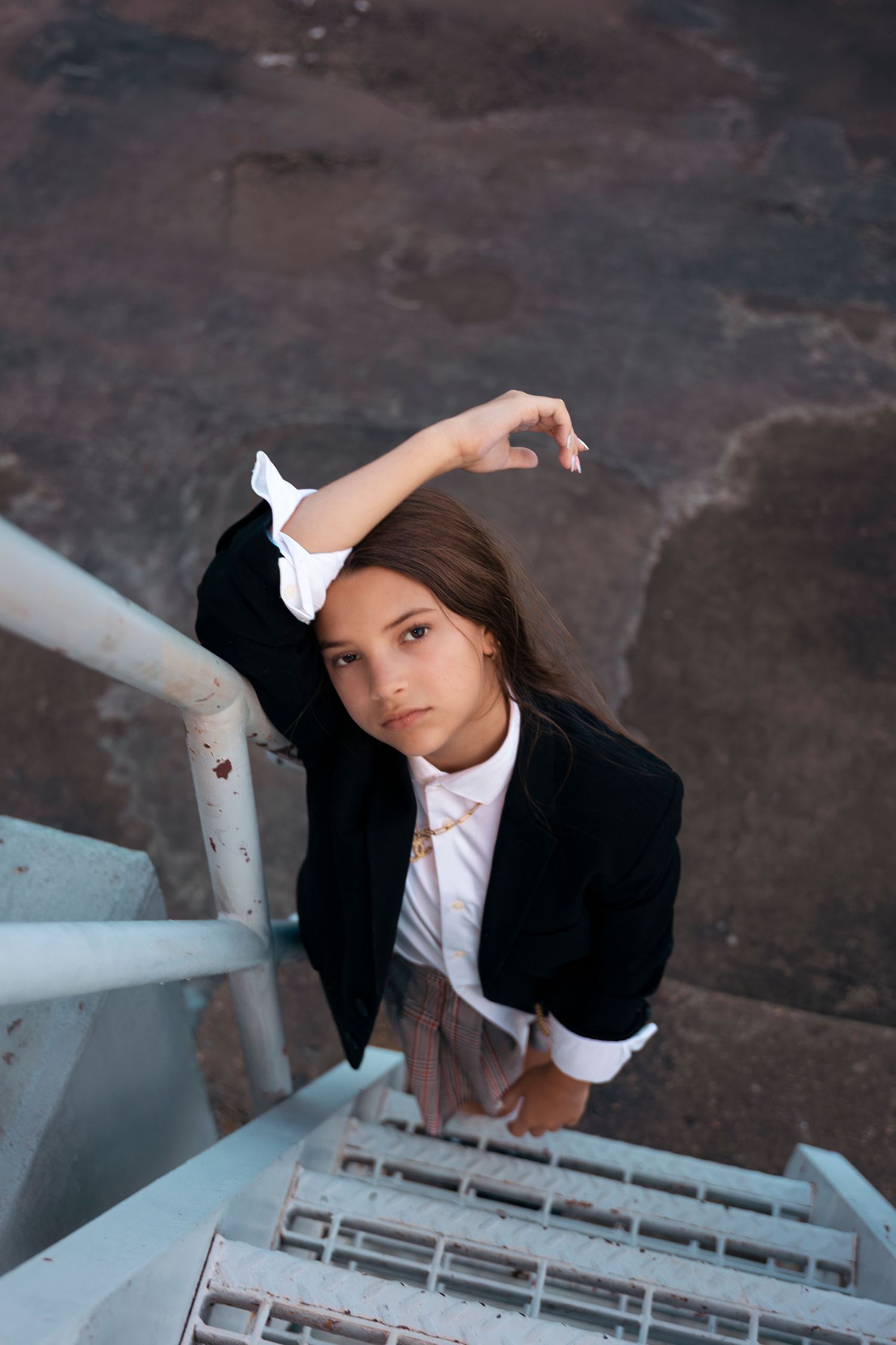 Young woman in black blazer and white shirt posing on metal staircase viewed from above against stone background.