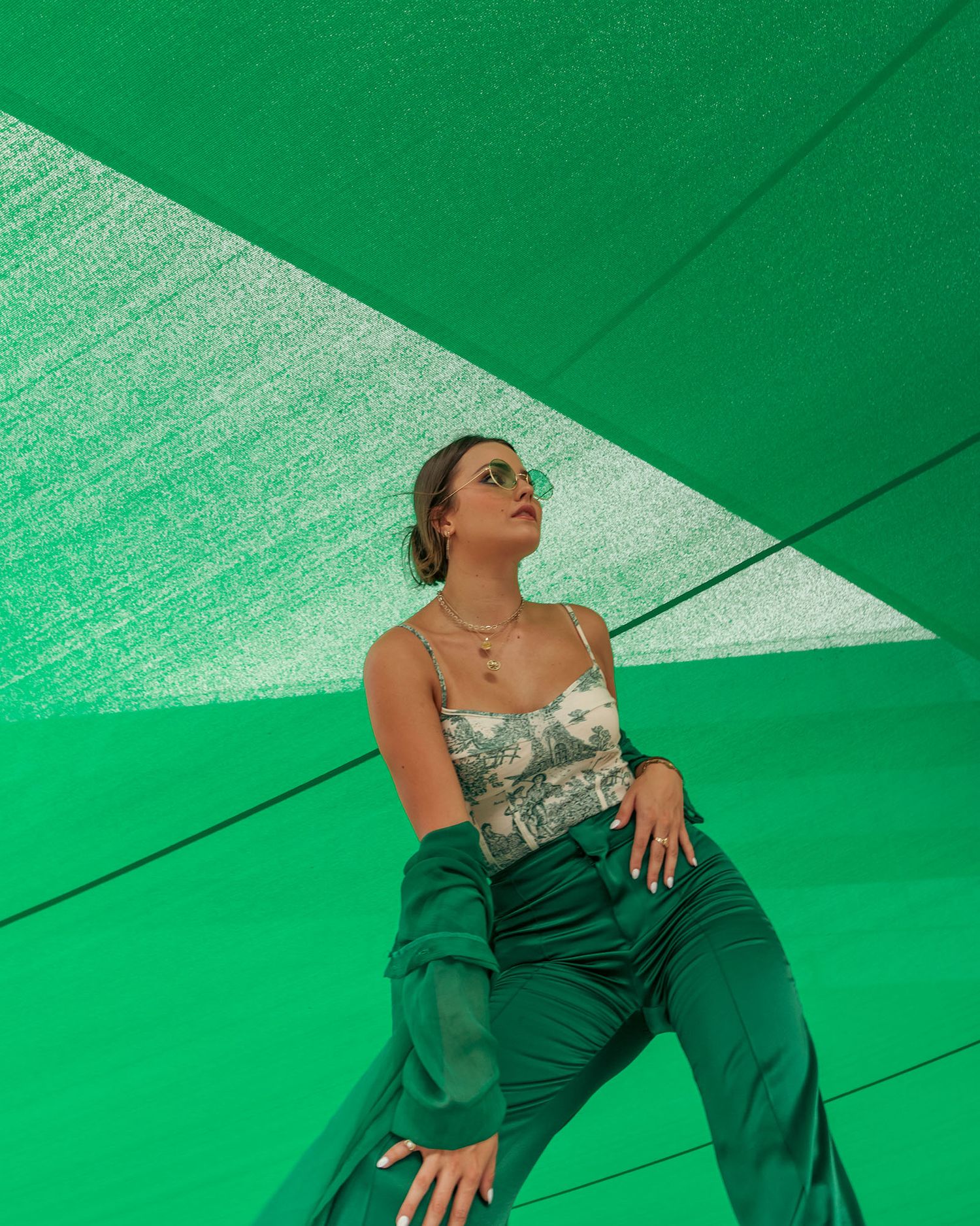 Woman in green wide-leg pants and floral top poses under vibrant green canopy structure.