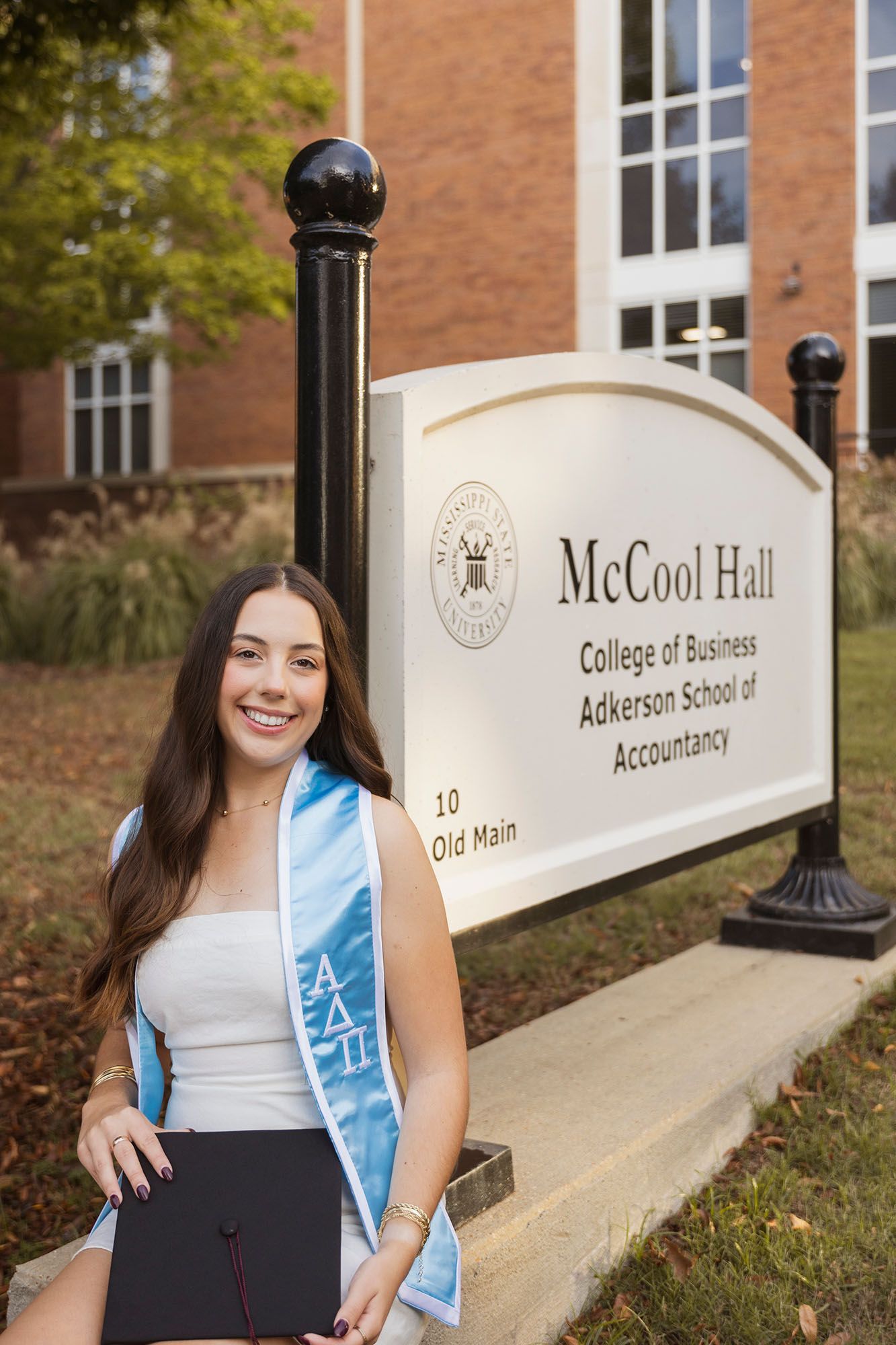 Smiling graduate in white dress holding cap beside McCool Hall College of Business sign.
