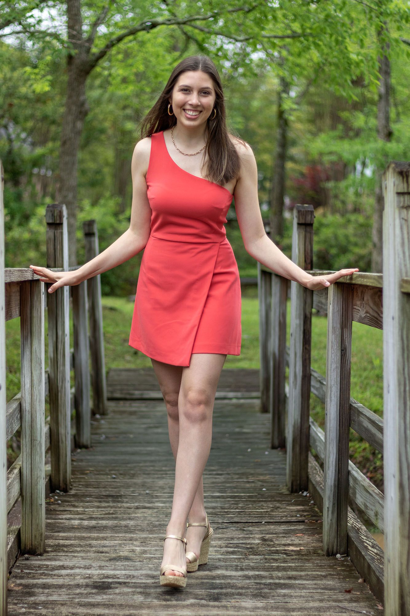Smiling young woman in red mini dress posing on wooden bridge in lush green park.