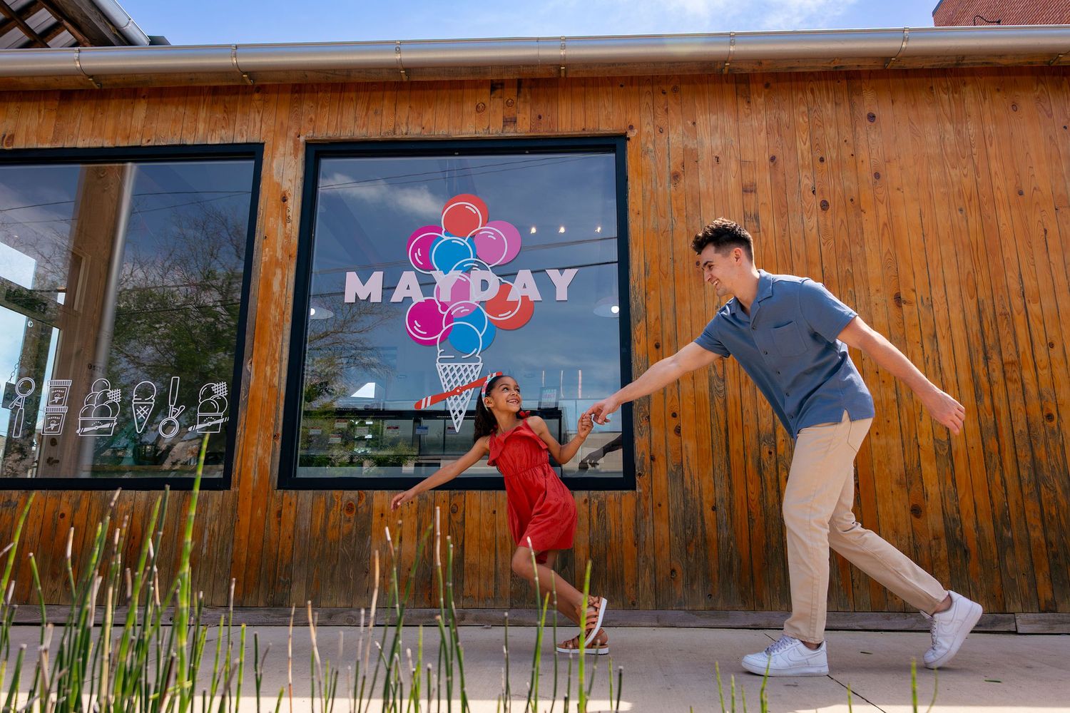 A young couple holds hands and plays outside a wooden building with a colorful Mayday sign.