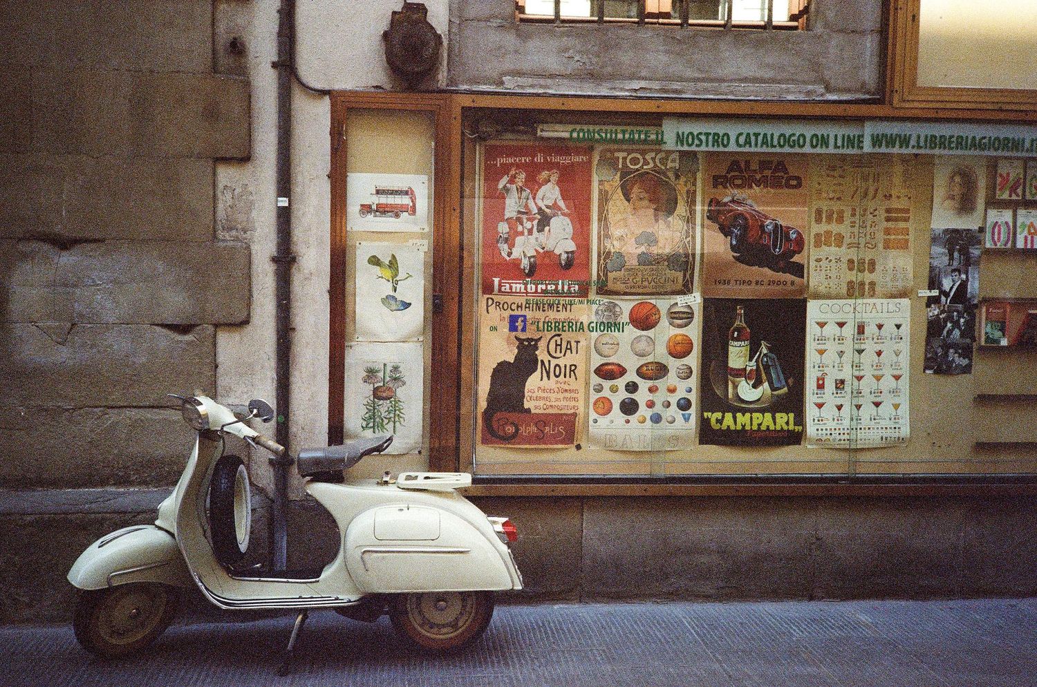 A vintage cream Vespa scooter parked outside a shop with colorful vintage posters in its window.