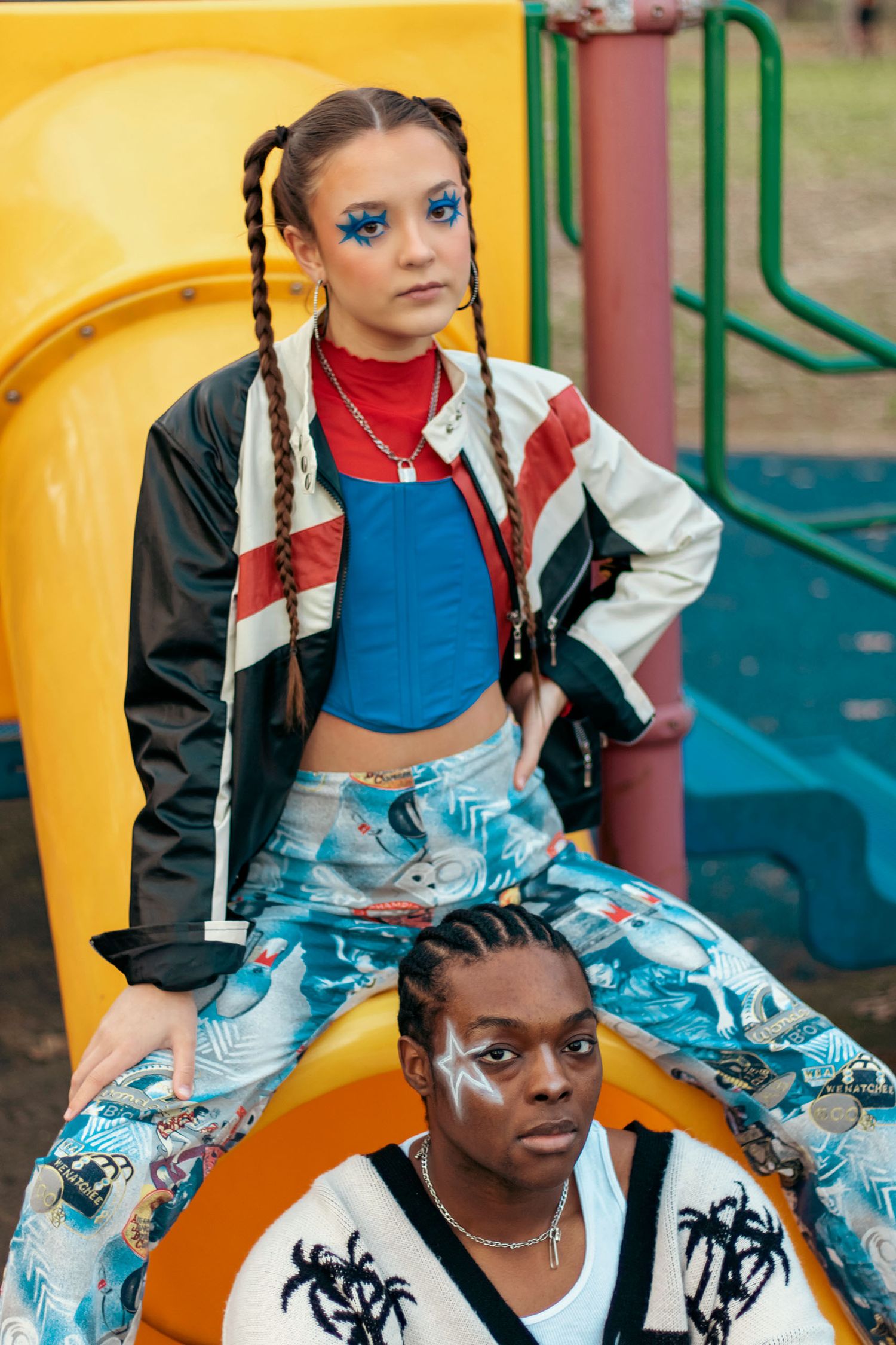 Two fashion models in streetwear posing at a colorful playground, woman in braids and blue eye makeup.
