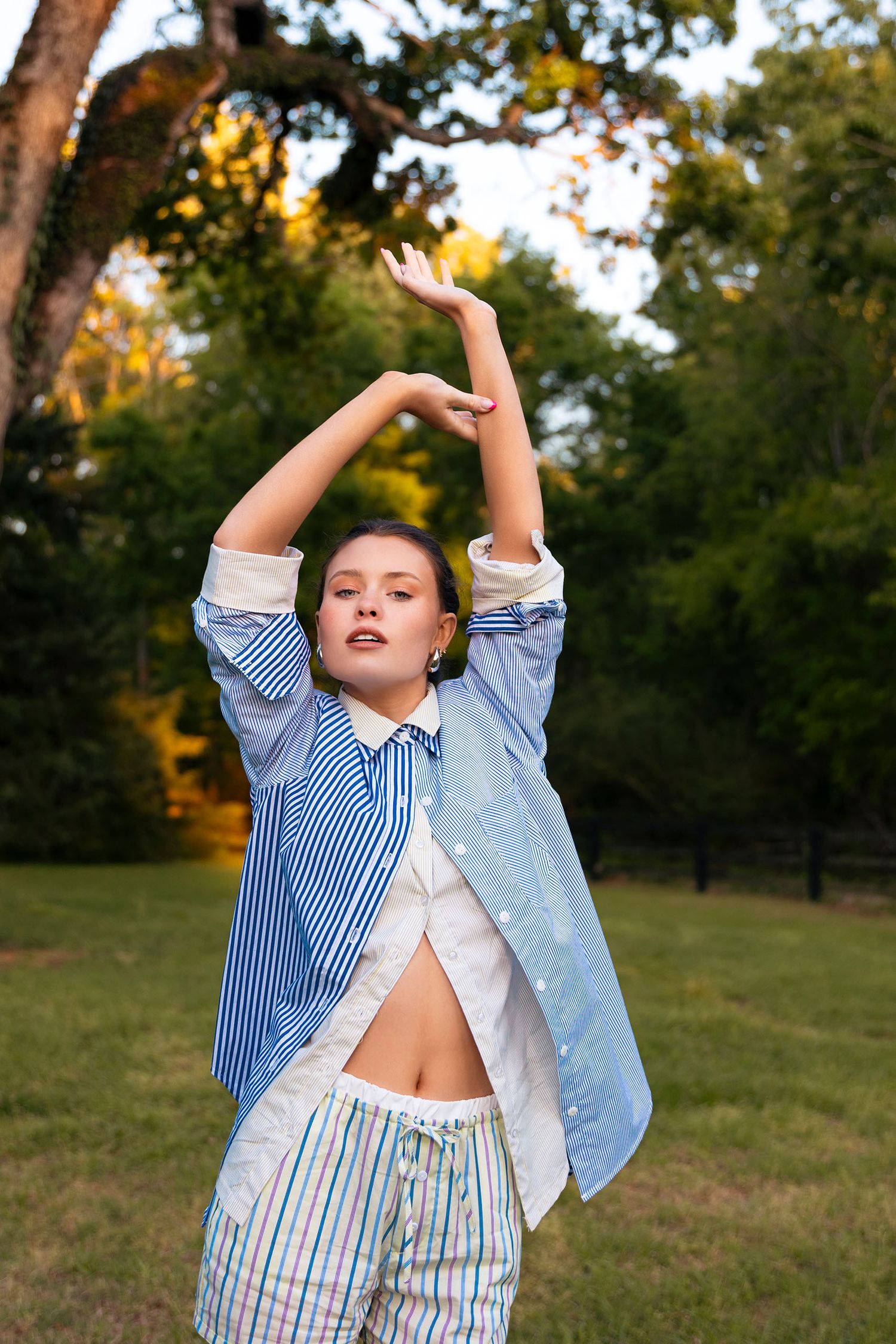 Young woman in blue striped shirt with arms raised posing in green outdoor setting.