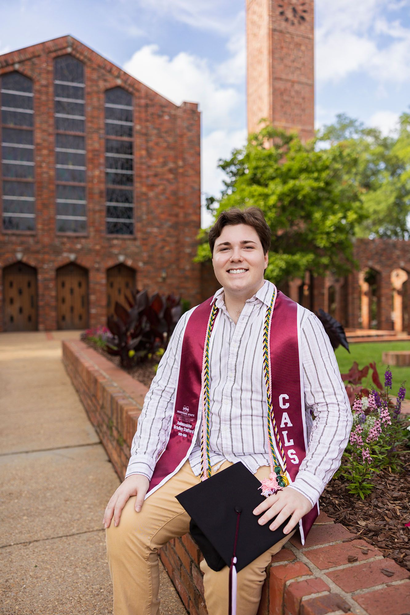 Smiling male graduate in striped shirt with maroon CALS stole sits outside brick chapel.