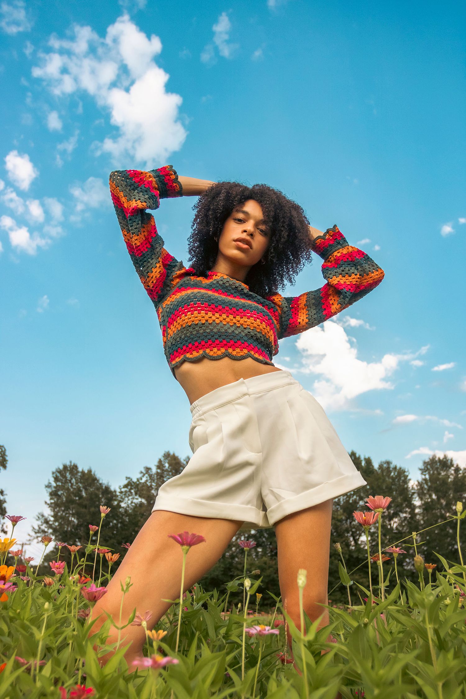 Woman in colorful crochet crop top and white shorts posing in a vibrant wildflower field outdoors.
