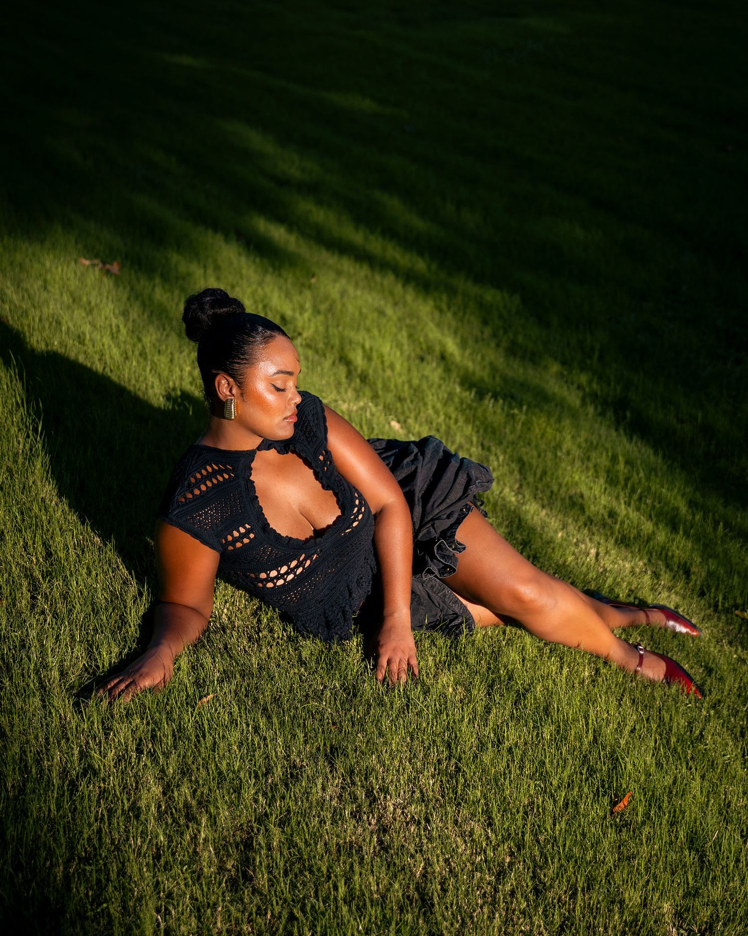 Woman in black crochet outfit reclines on green grass in dramatic sunlight.