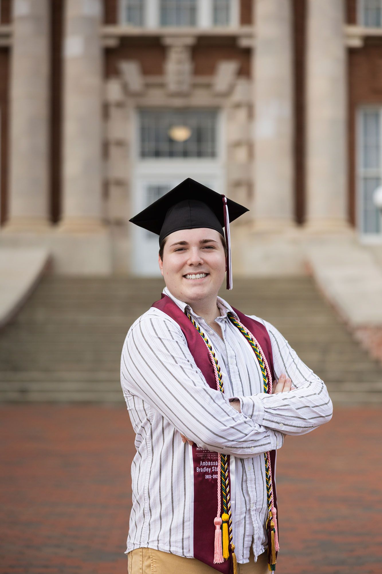 Male graduate smiles confidently in cap and honor stoles, arms crossed before historic university building.