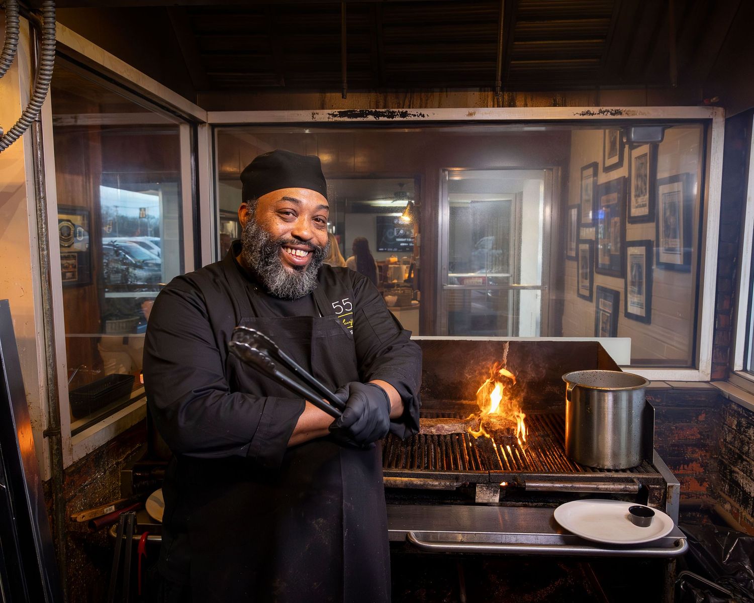 Smiling chef grilling meat over open flames at an outdoor BBQ station with tongs in hand.