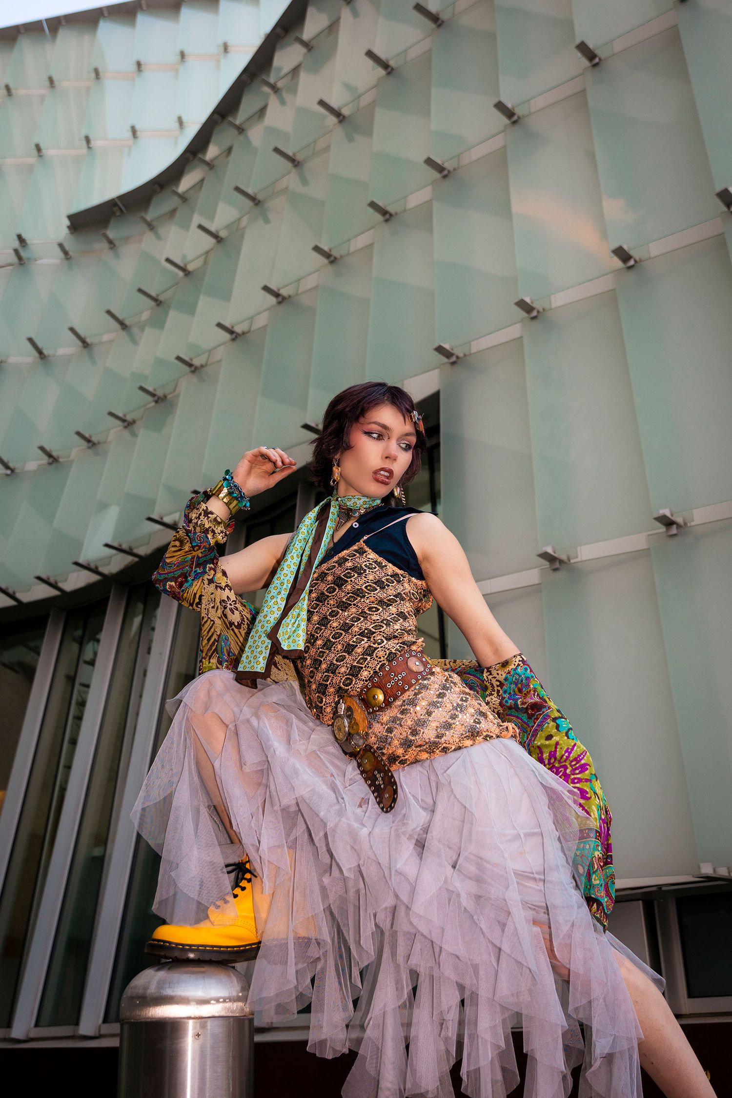 Model in eclectic mixed-print outfit with tulle skirt and yellow boots near modern building.