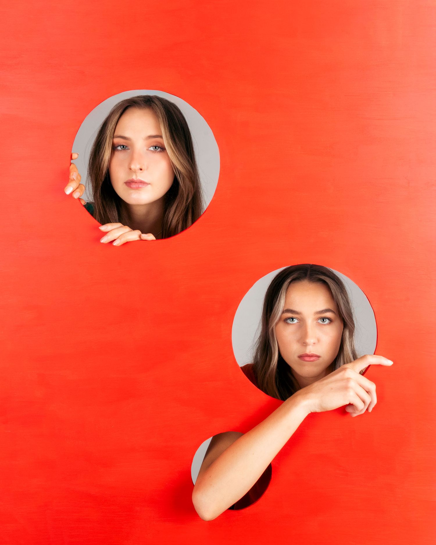 Two young women peek through circular holes in bright red paper background.
