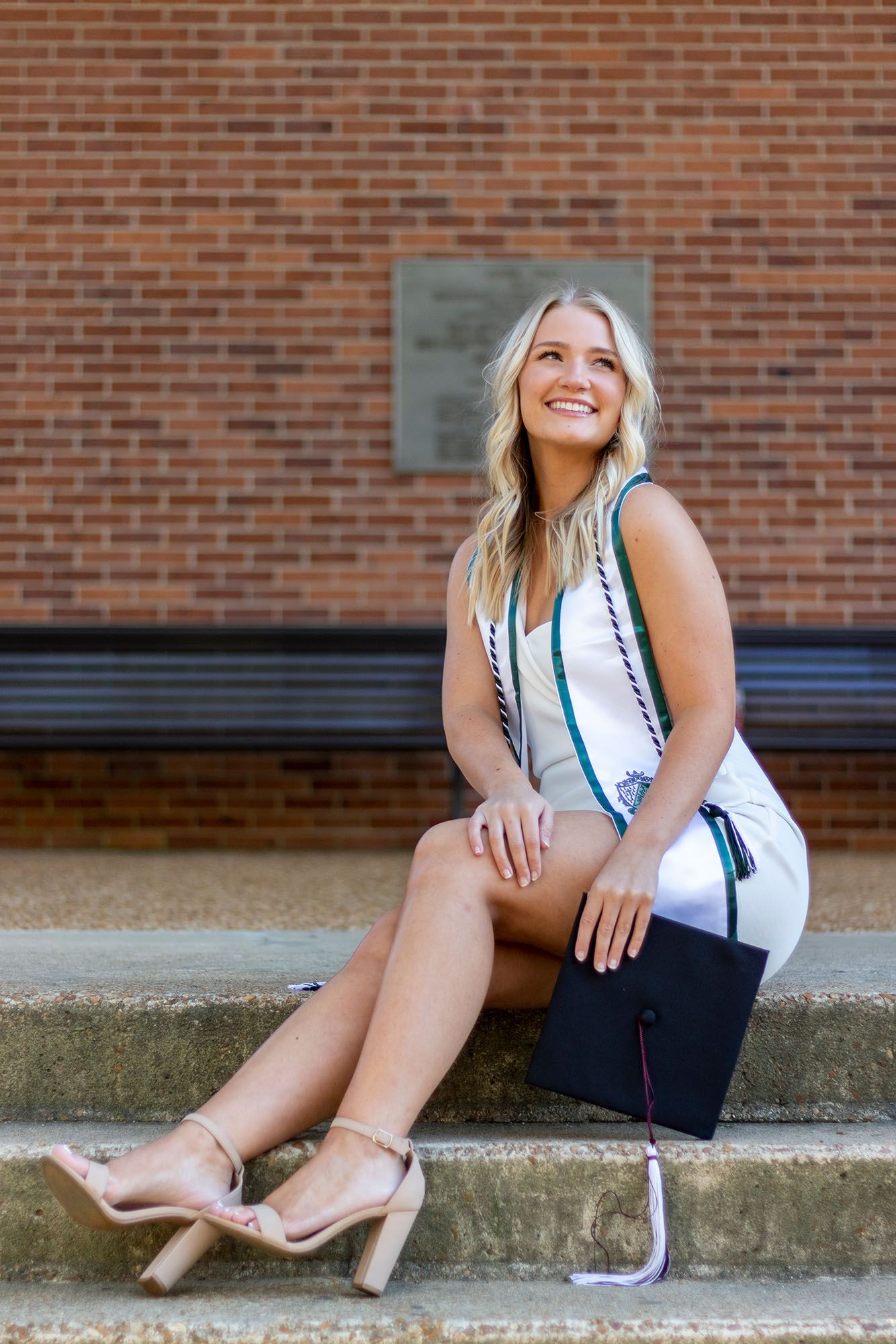 Smiling blonde graduate sits on steps holding cap, wearing white dress with honor cords and heels against brick wall.