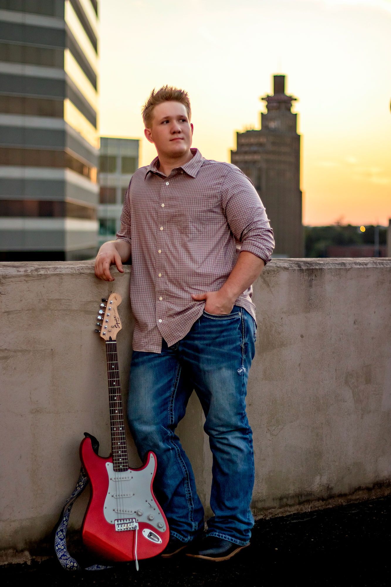 Young man leaning on red electric guitar at rooftop with city skyline at sunset.