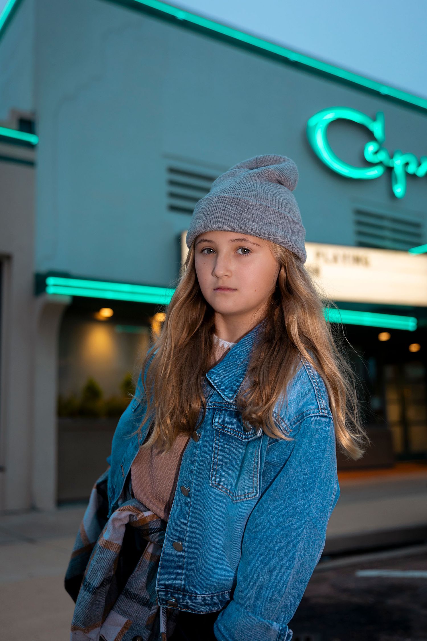 A teenage girl in a denim jacket and gray beanie stands outside a cinema at dusk.
