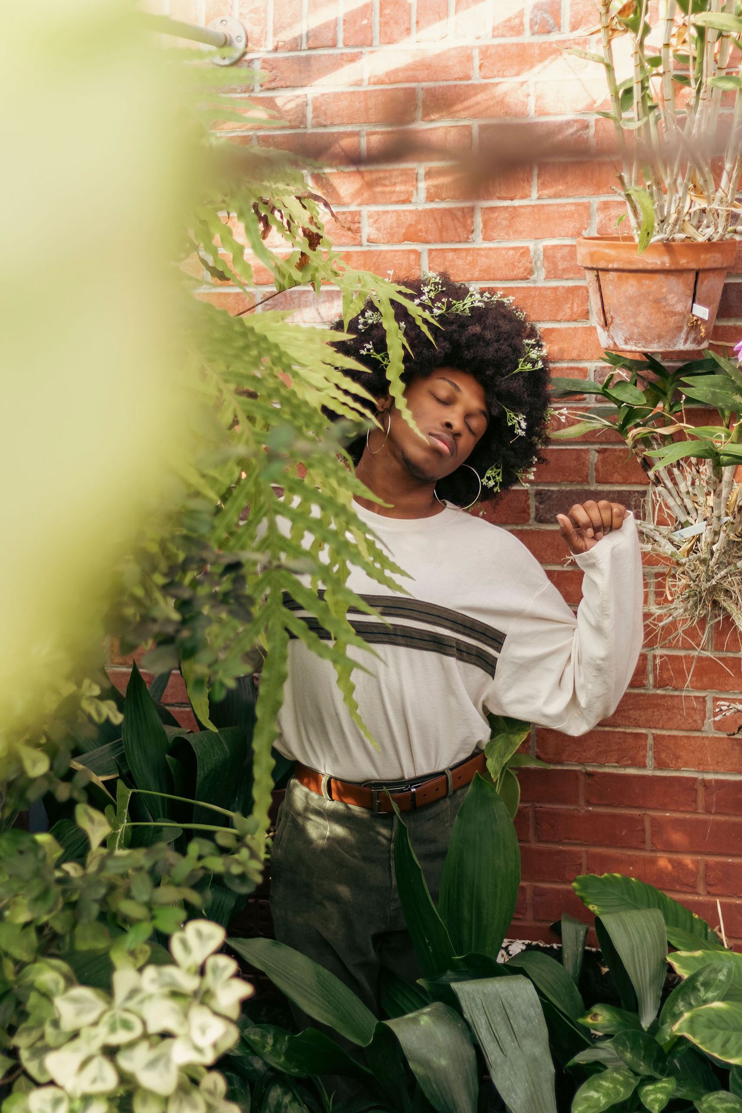 A woman with an afro poses among lush green tropical plants against a brick wall.